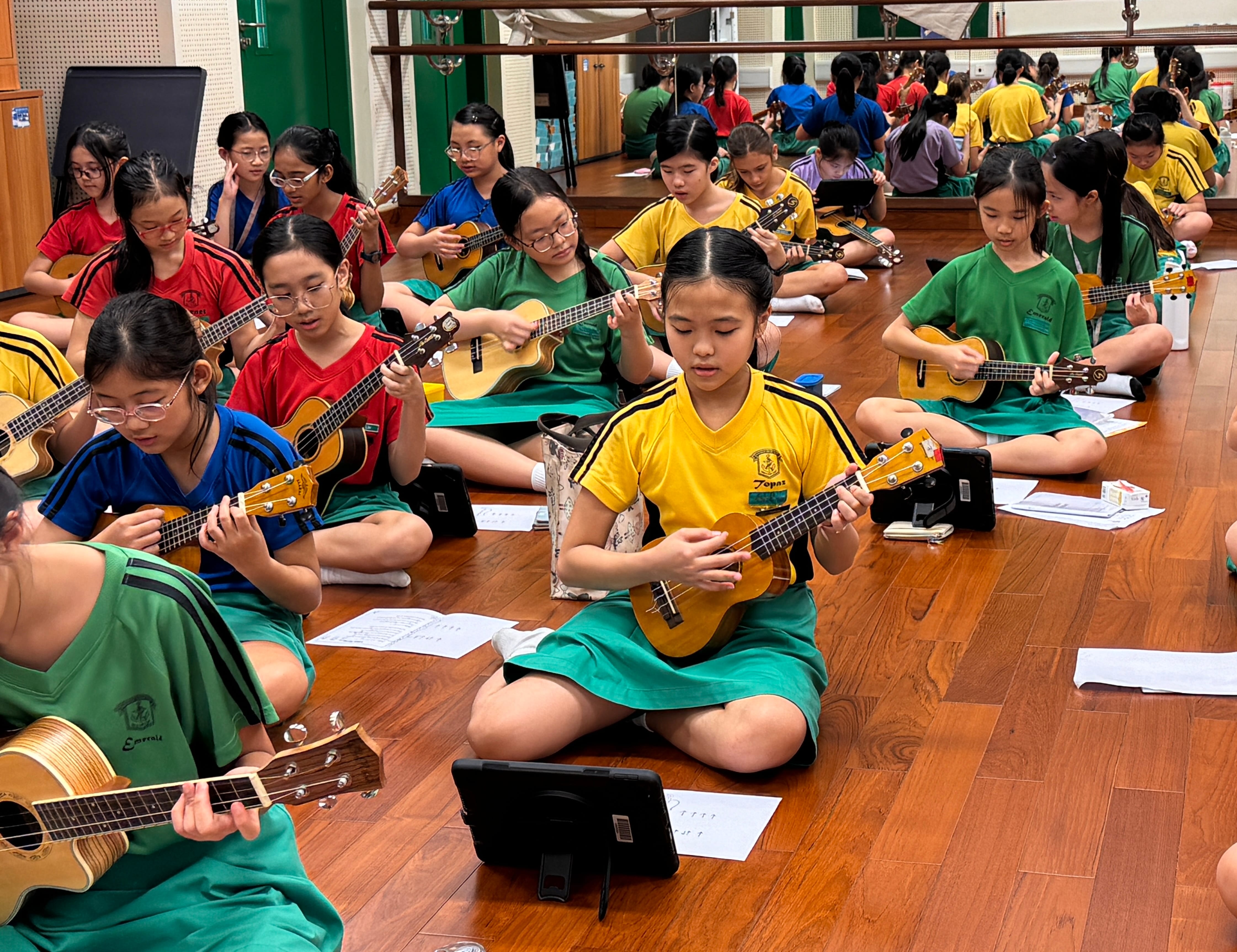 Many school children in colorful uniforms sit cross-legged on a wooden floor playing ukuleles in a large room.