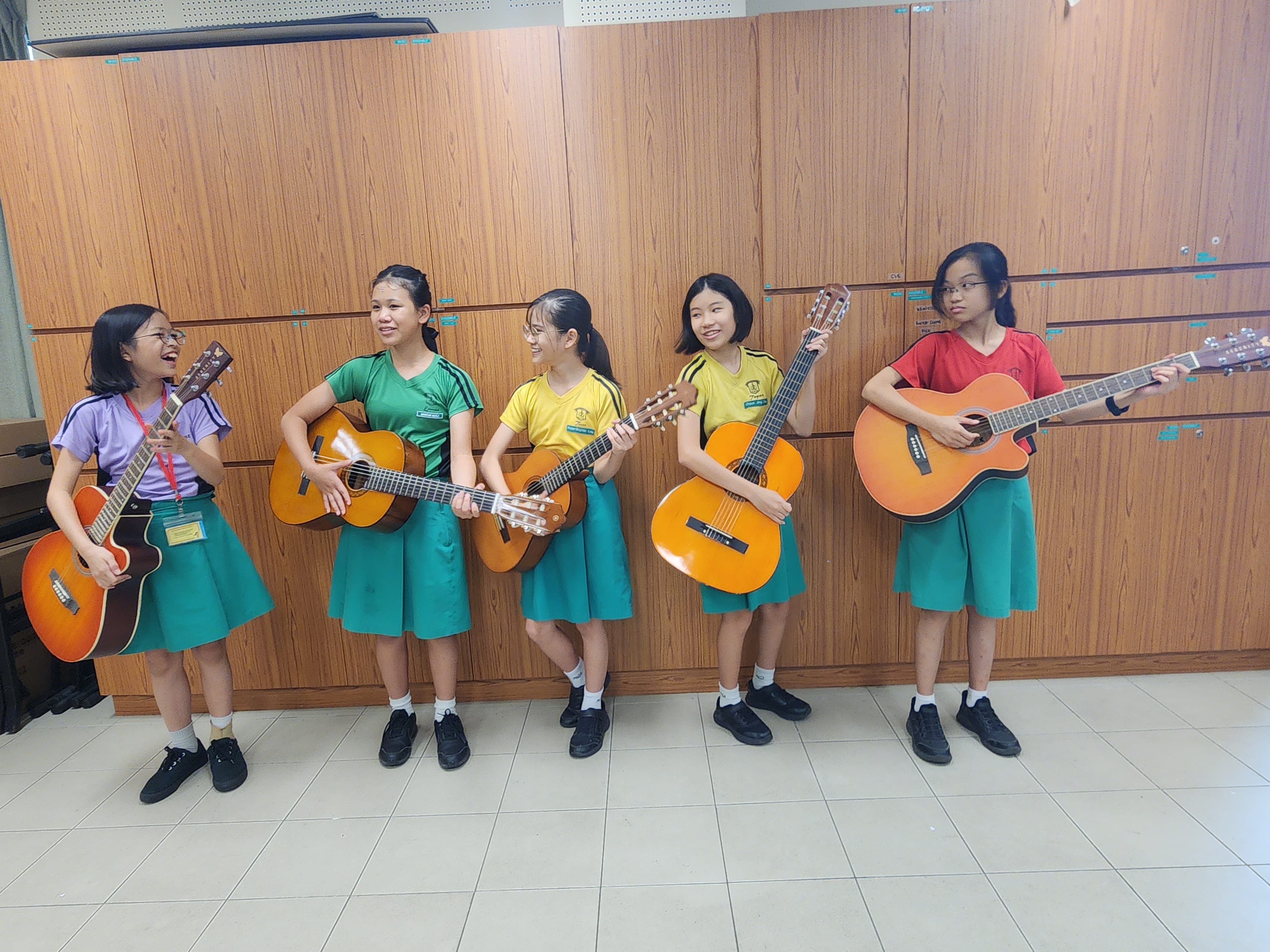 Five young female students in school uniforms hold acoustic guitars while standing in front of wooden lockers.