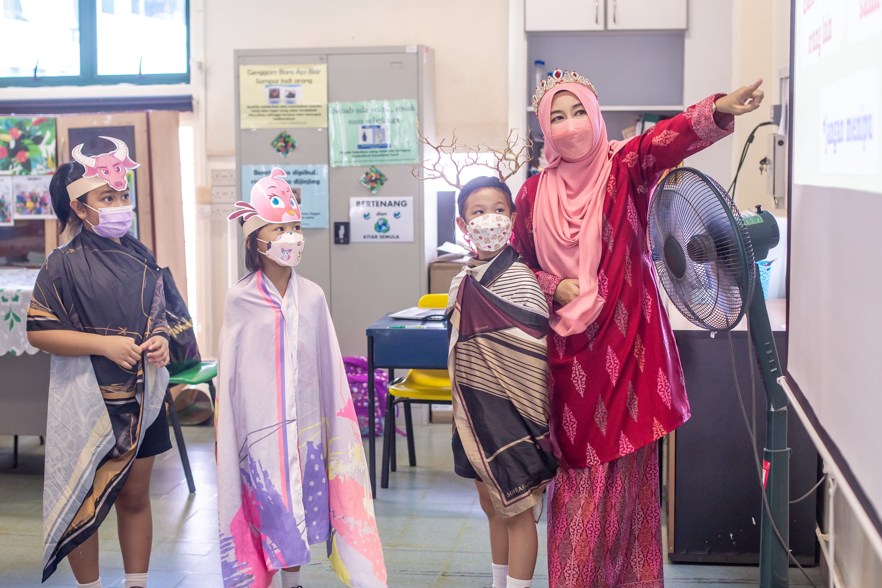 A teacher in a crown and red dress points at a board while three masked children wear costumes in a classroom.