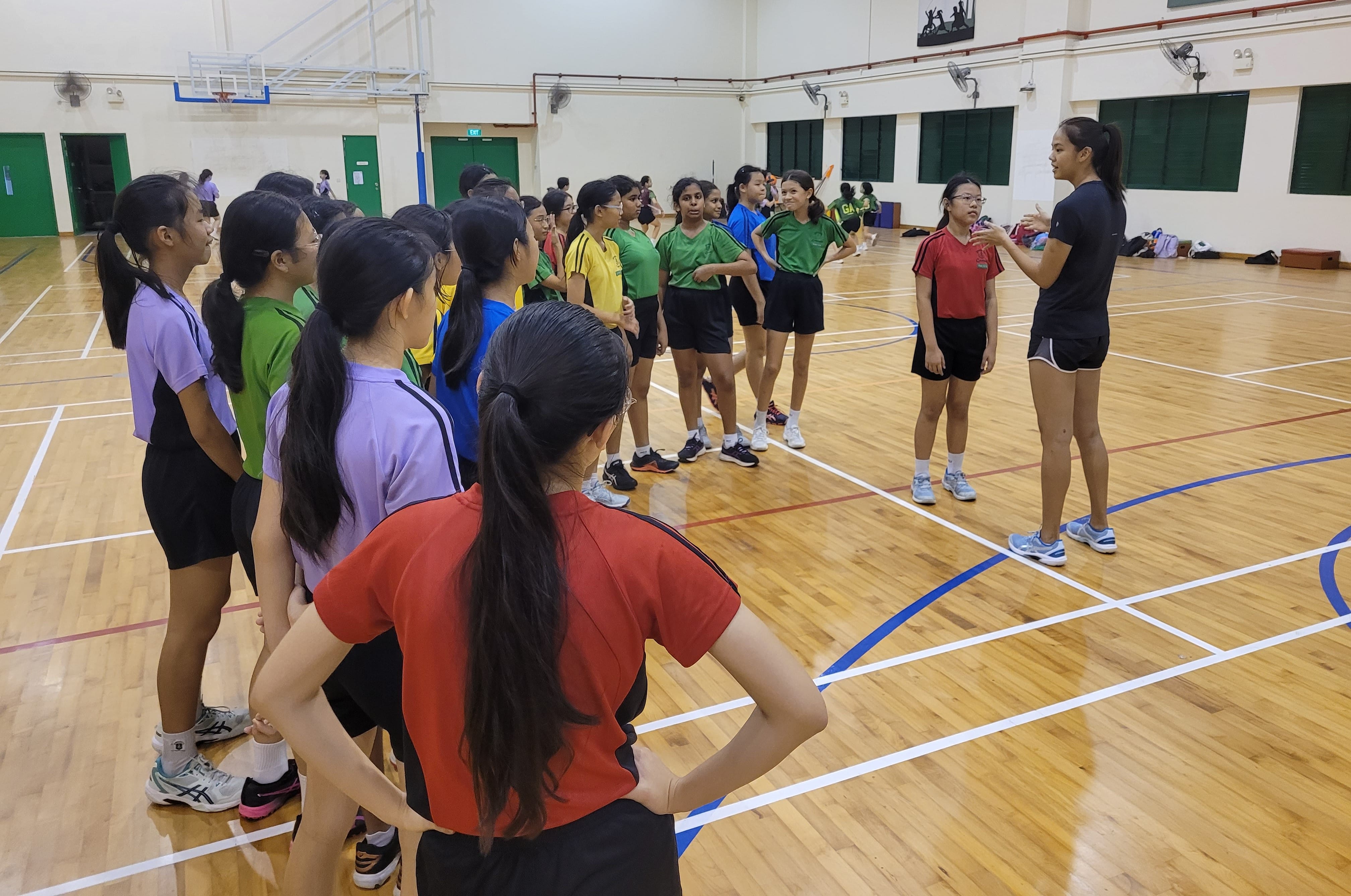 A group of young girls in colorful sportswear listen to a coach speaking on an indoor wooden court.