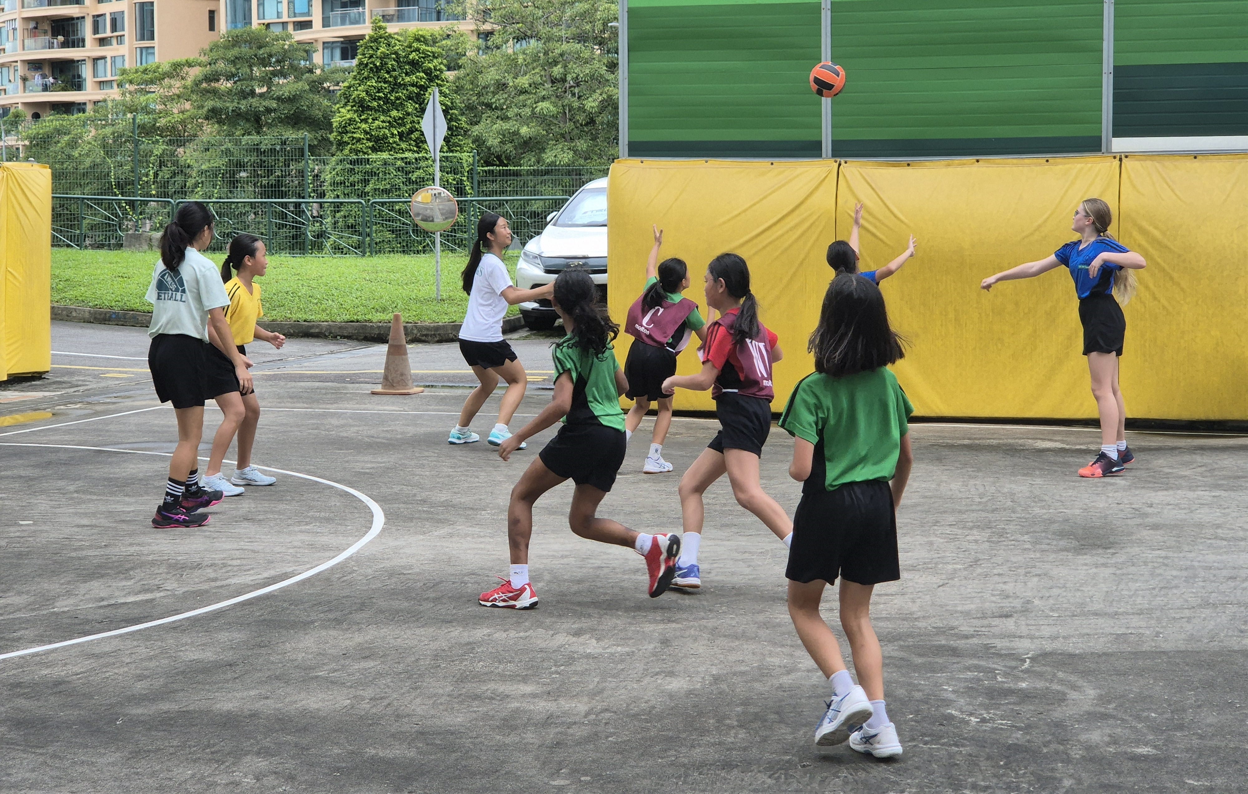Girls in green, red, and white jerseys play netball on an outdoor court with a ball in the air.