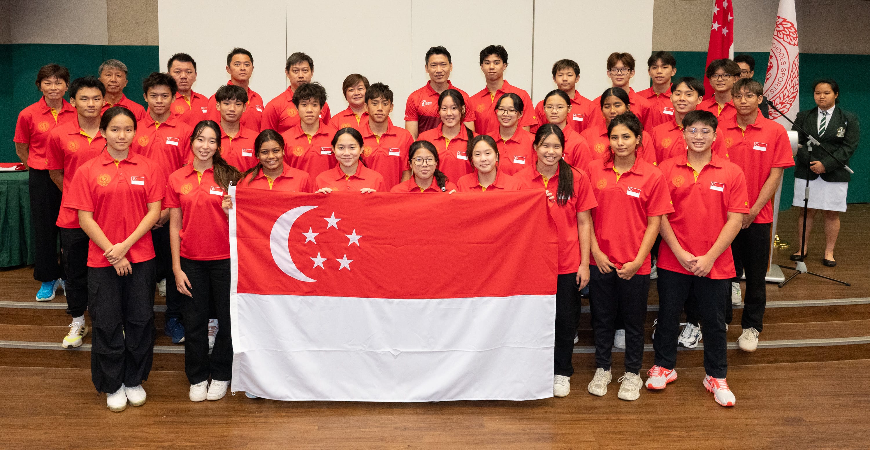 A large group in red Singapore team uniforms poses indoors holding the Singapore national flag.