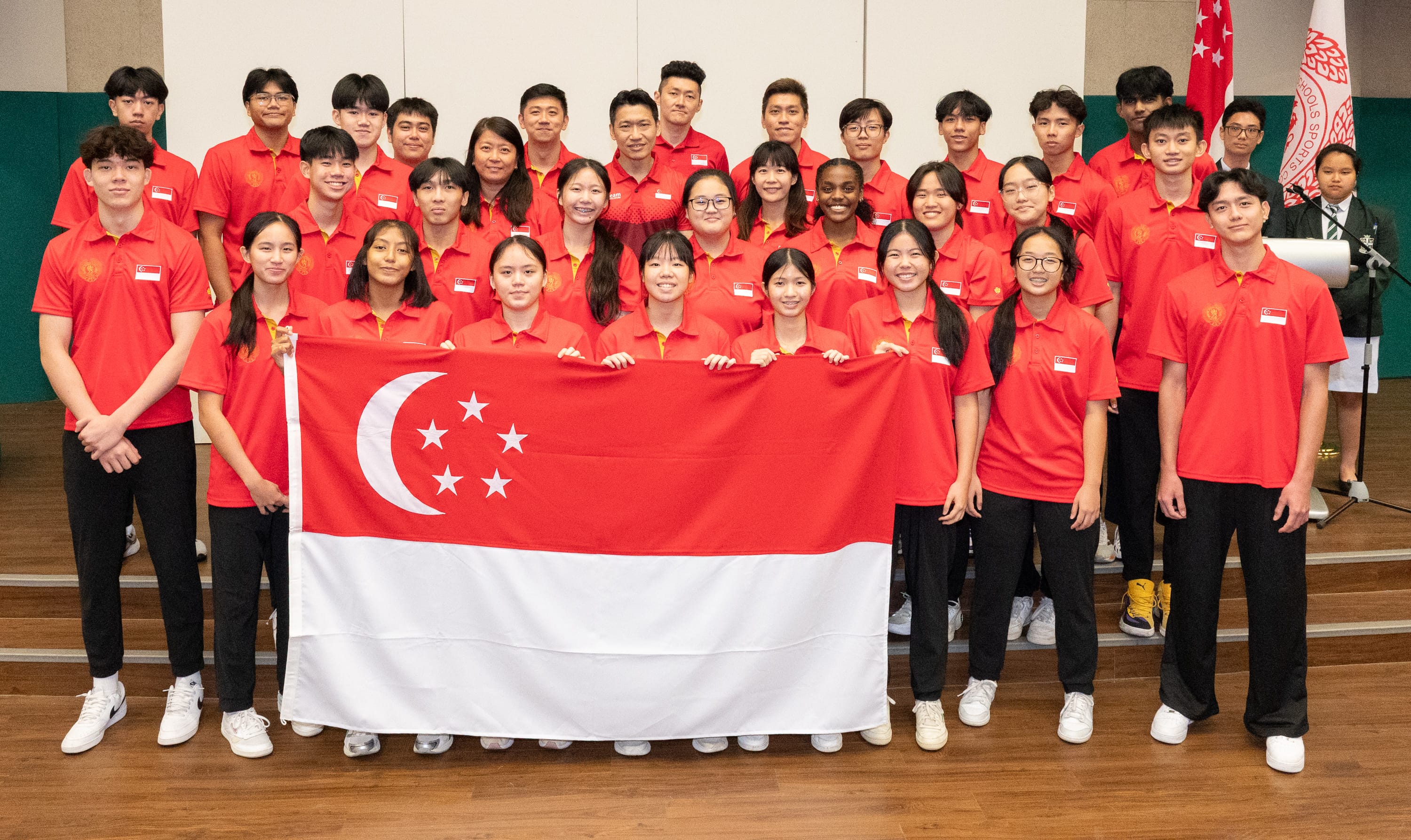 A large group of young people in red Singapore team polo shirts pose together holding the Singapore national flag.