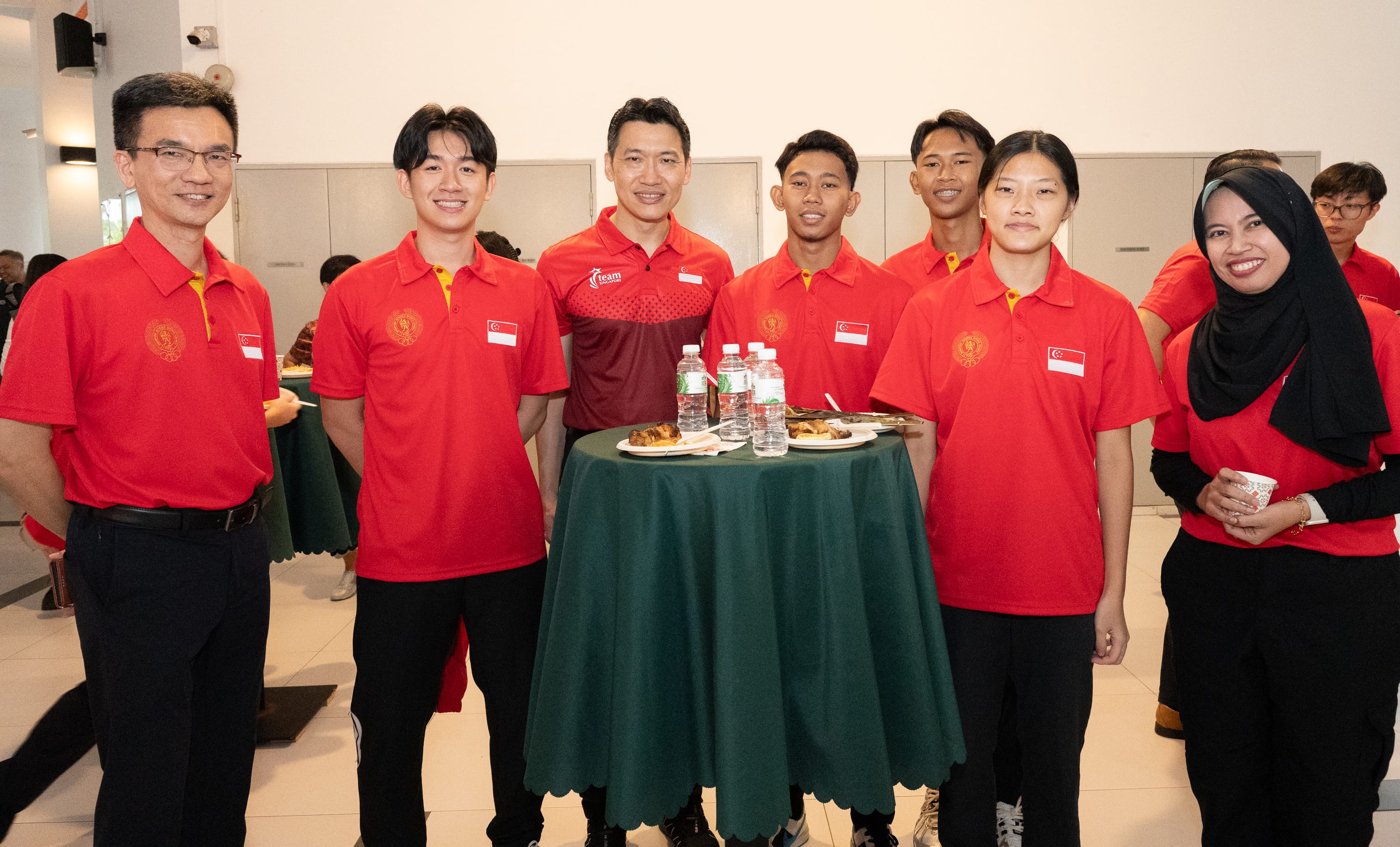 Seven people in red Singapore team polo shirts smile together around a green-clothed high table with food and water bottles.
