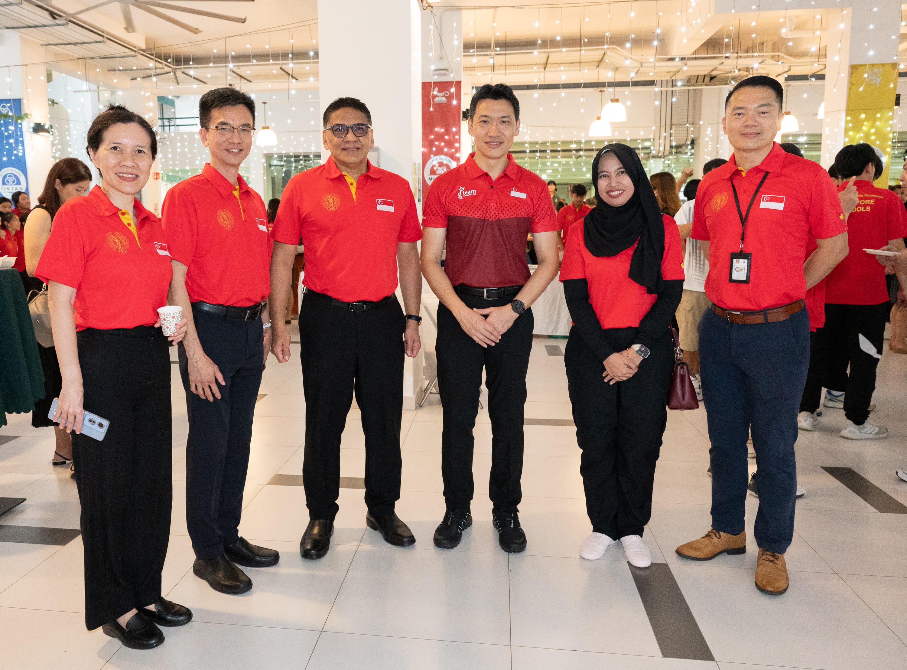 Six people in red polo shirts smile together indoors at a bright event venue with string lights overhead.