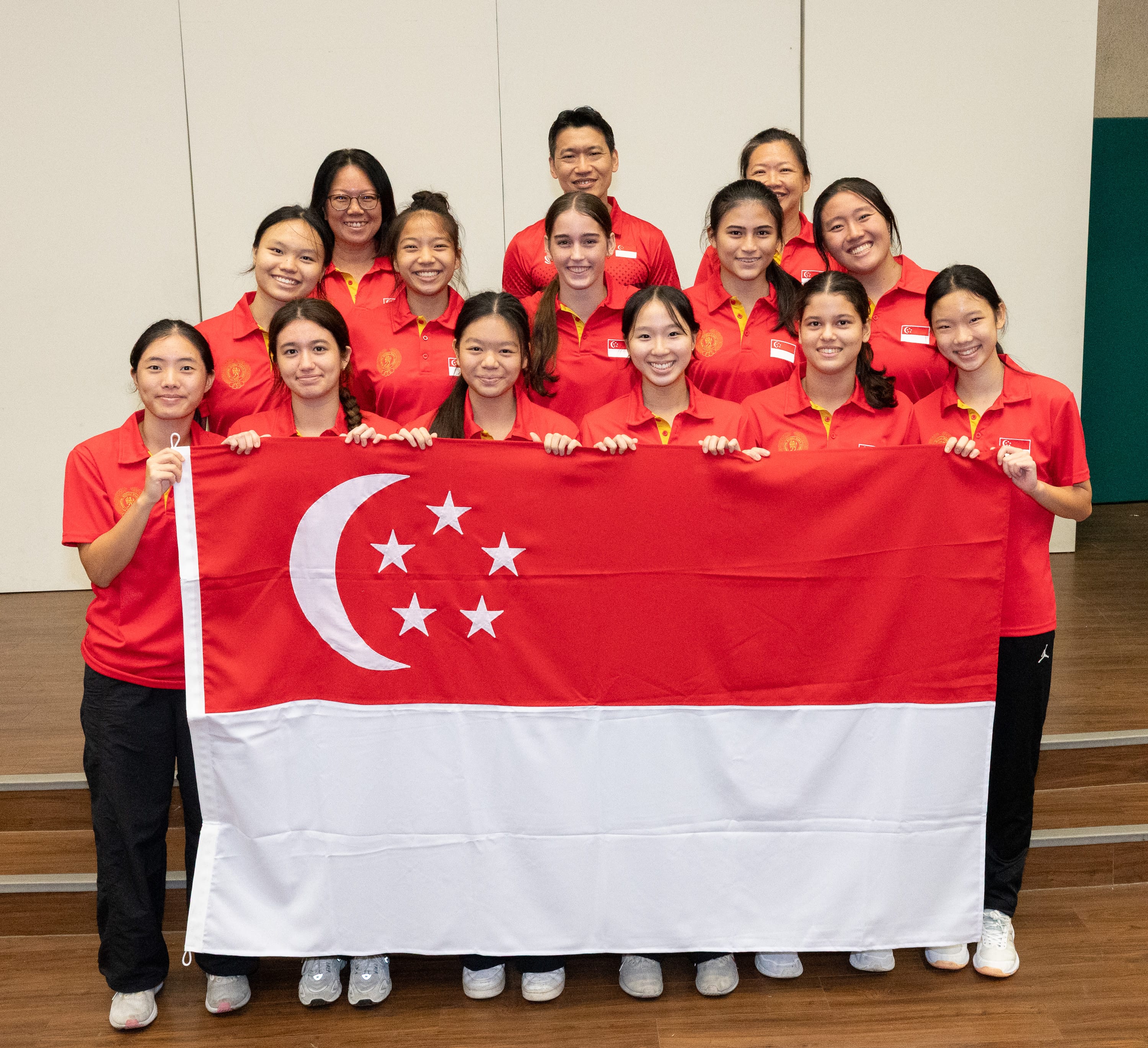 A group of smiling people in red Singapore team jerseys pose together holding the Singapore national flag.