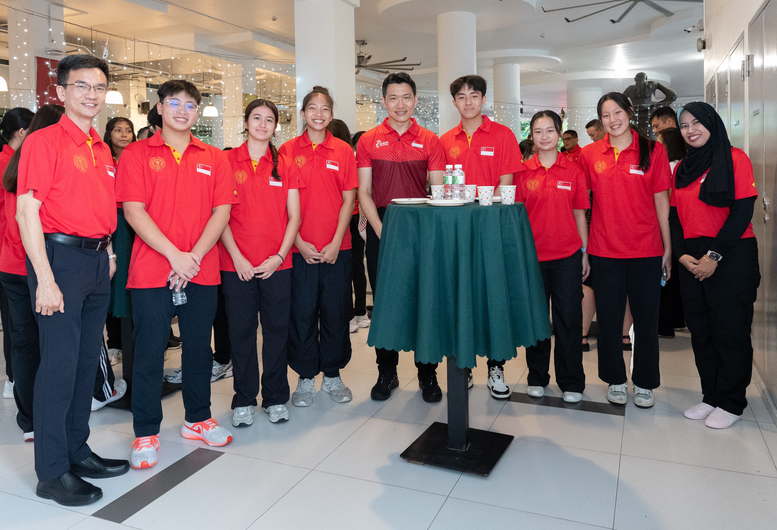 A group of people in matching red polo shirts with Singapore flags pose together around a green cocktail table indoors.