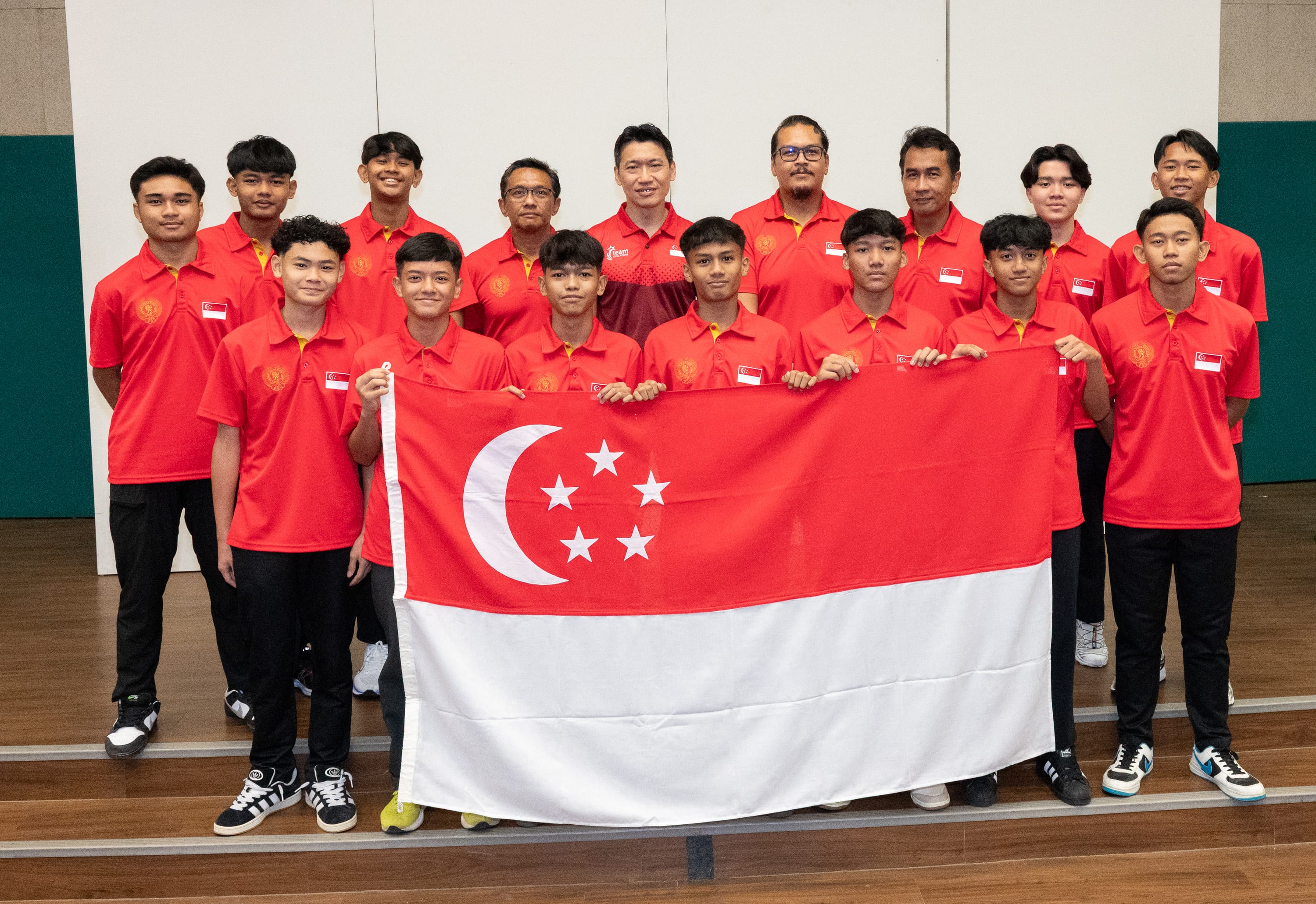 A group of young males in red Singapore team polo shirts pose together holding the Singapore national flag indoors.