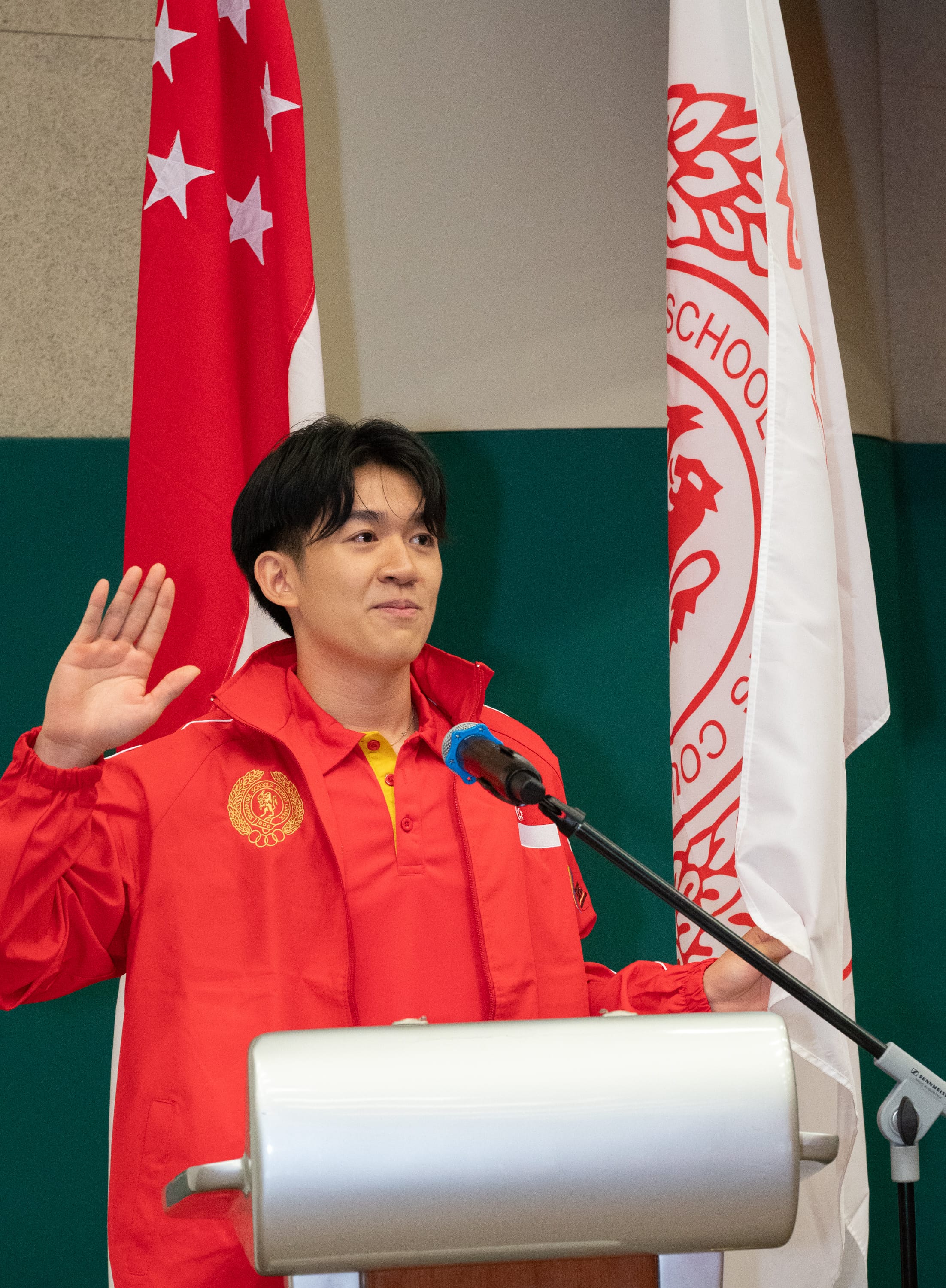 A young man in a red jacket raises his right hand at a podium with the Singapore flag behind him.