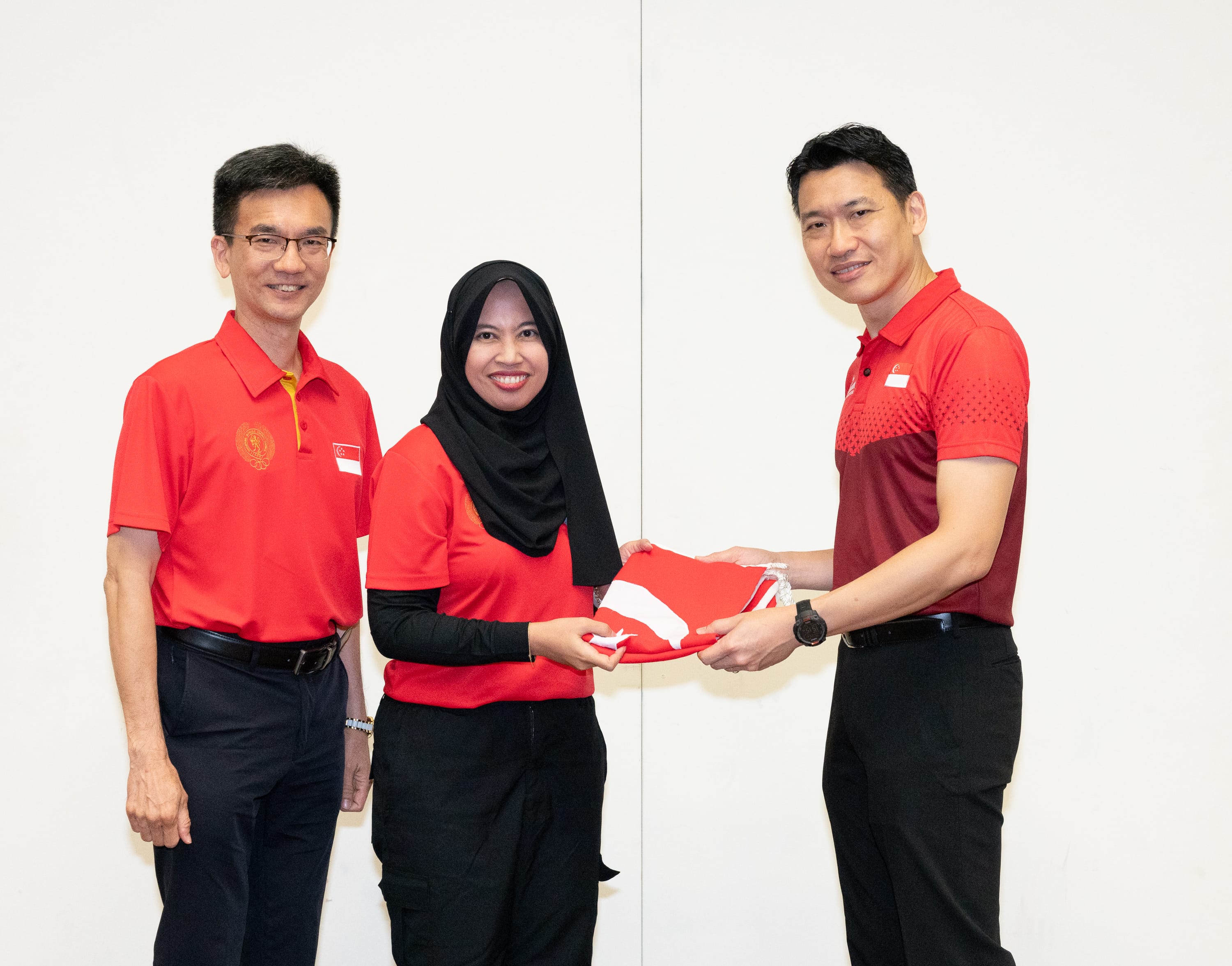 Three people in red shirts; two hold a folded Singapore flag while the third stands beside them.