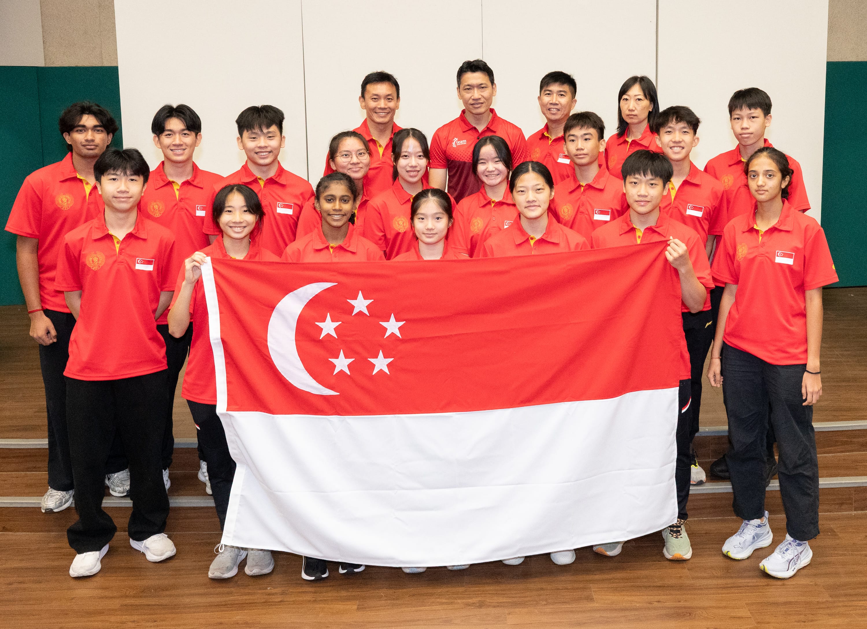 A group of young athletes in red Singapore team jerseys posing together while holding the Singapore national flag.
