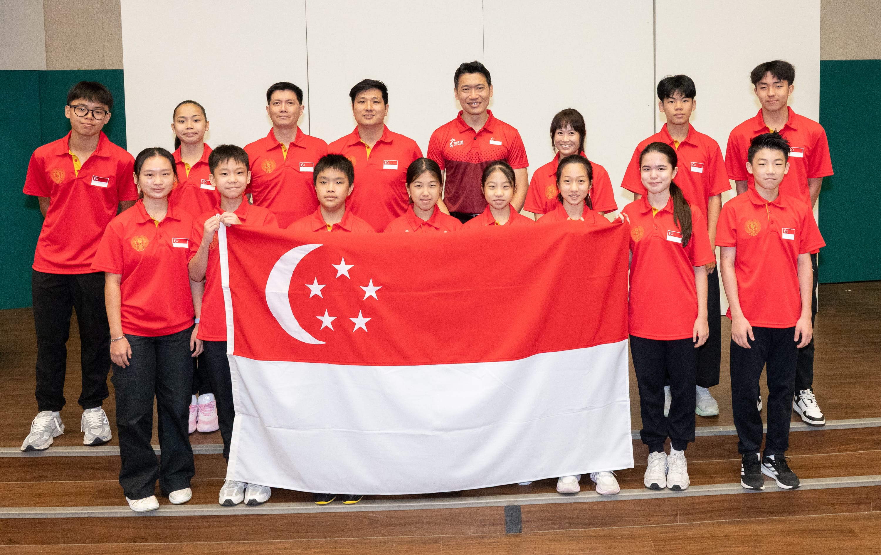 A group of young athletes and coaches in red Singapore jerseys pose together holding the Singapore national flag.
