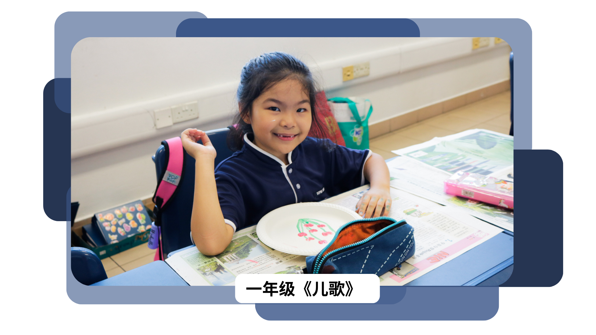 A smiling young girl in a navy school uniform sits at a desk with a painted plate and pencil case.