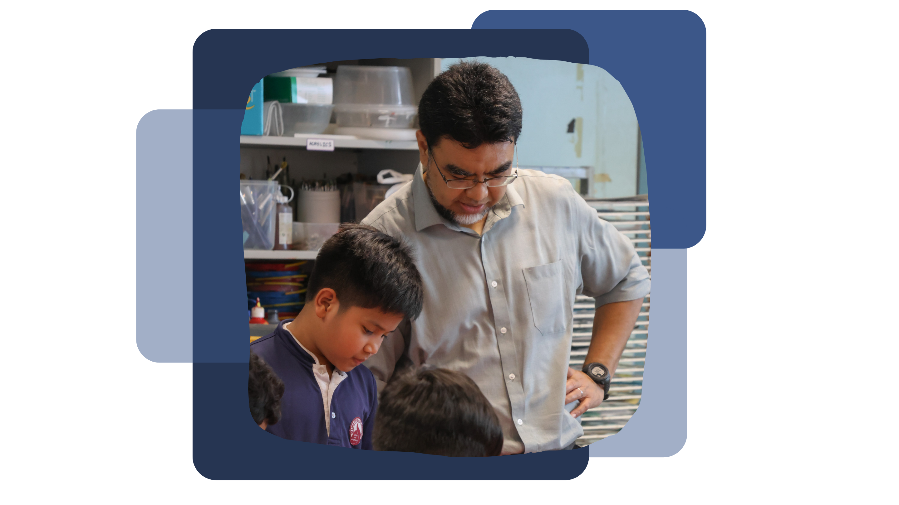 A male teacher with glasses leans over two male students in school uniforms inside an art room.