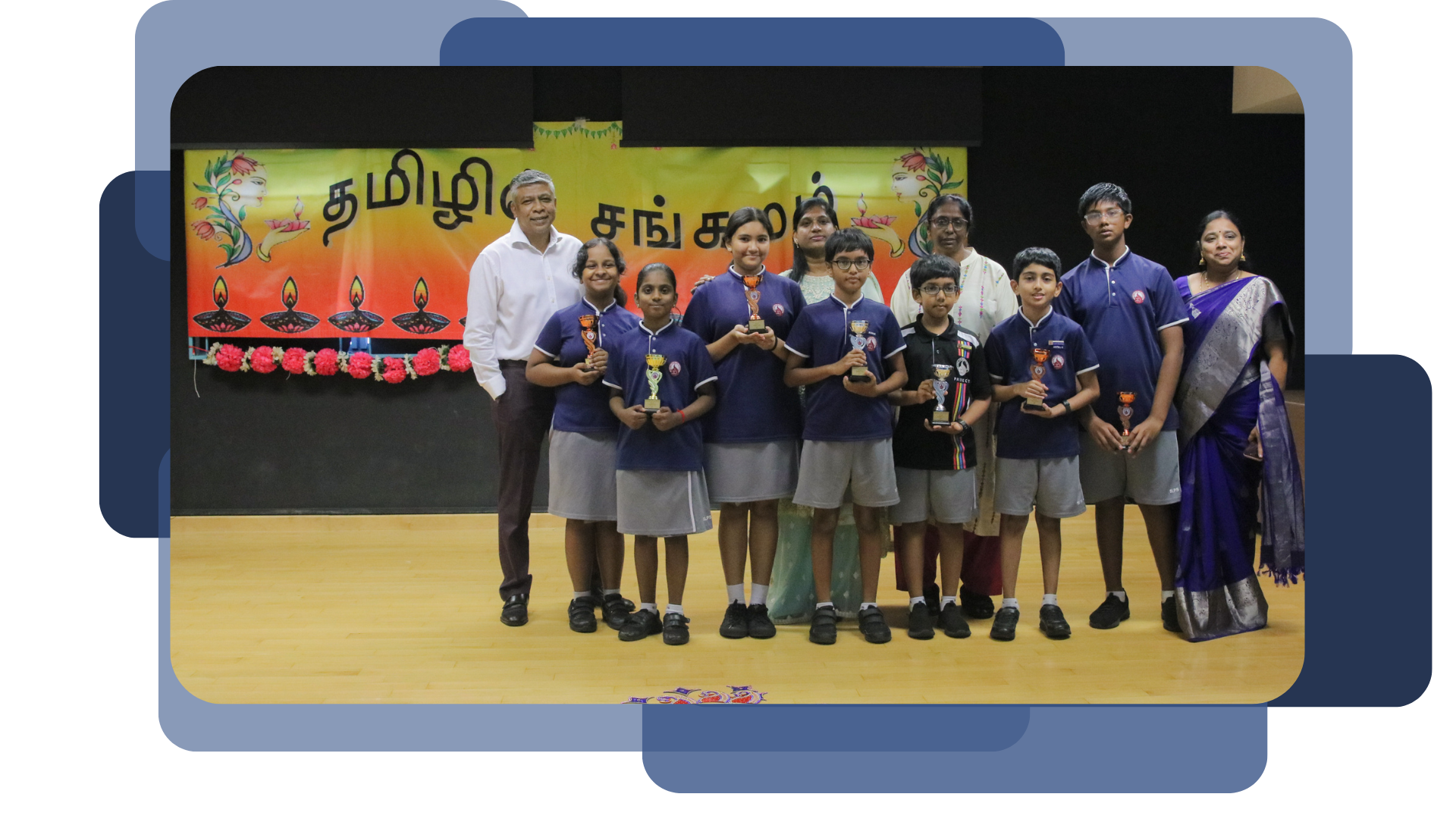 Students in blue uniforms hold trophies on a stage in front of a Tamil-script banner with adults beside them.