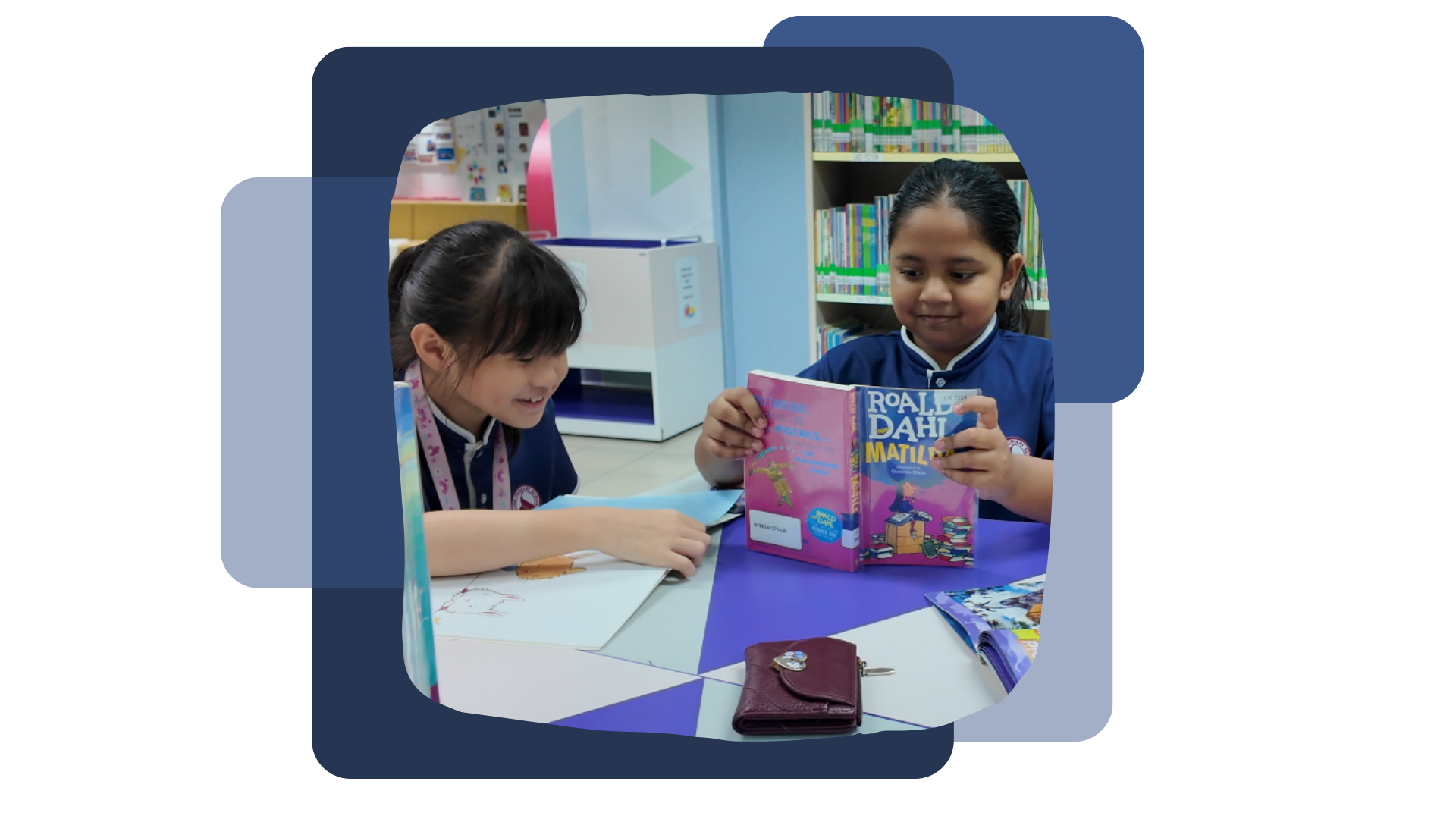 Two young girls in school uniforms sit at a library table, one holding Roald Dahl's Matilda book.