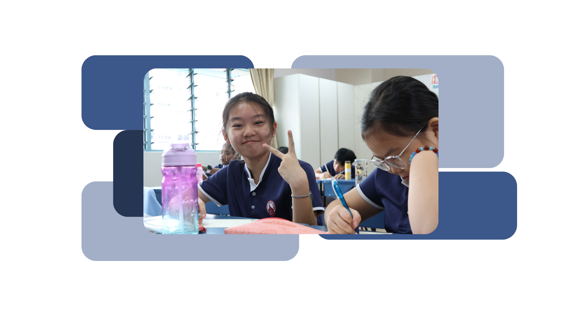 Two young girls in navy school uniforms sit at classroom desks, one smiling and making a peace sign.