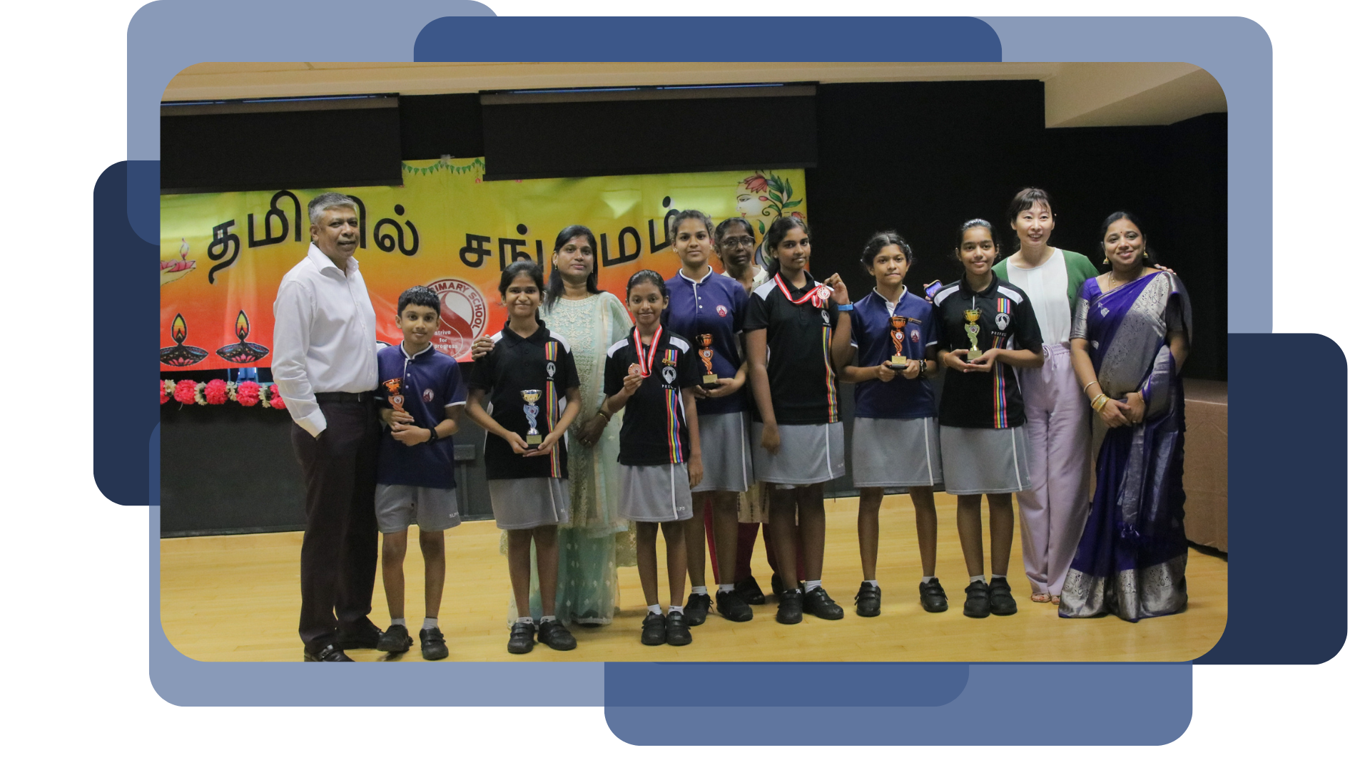 Students in uniforms holding trophies and medals stand with adults on a stage in front of a Tamil banner.