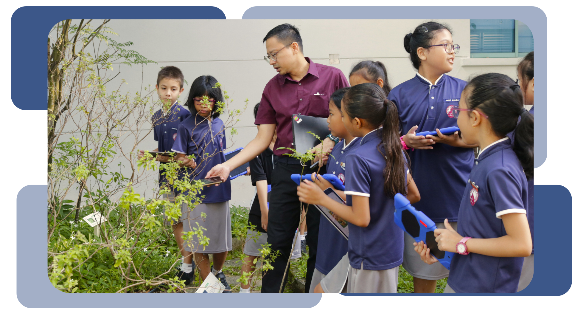A teacher in a maroon shirt guides a group of students in blue uniforms examining outdoor plants with tablets.
