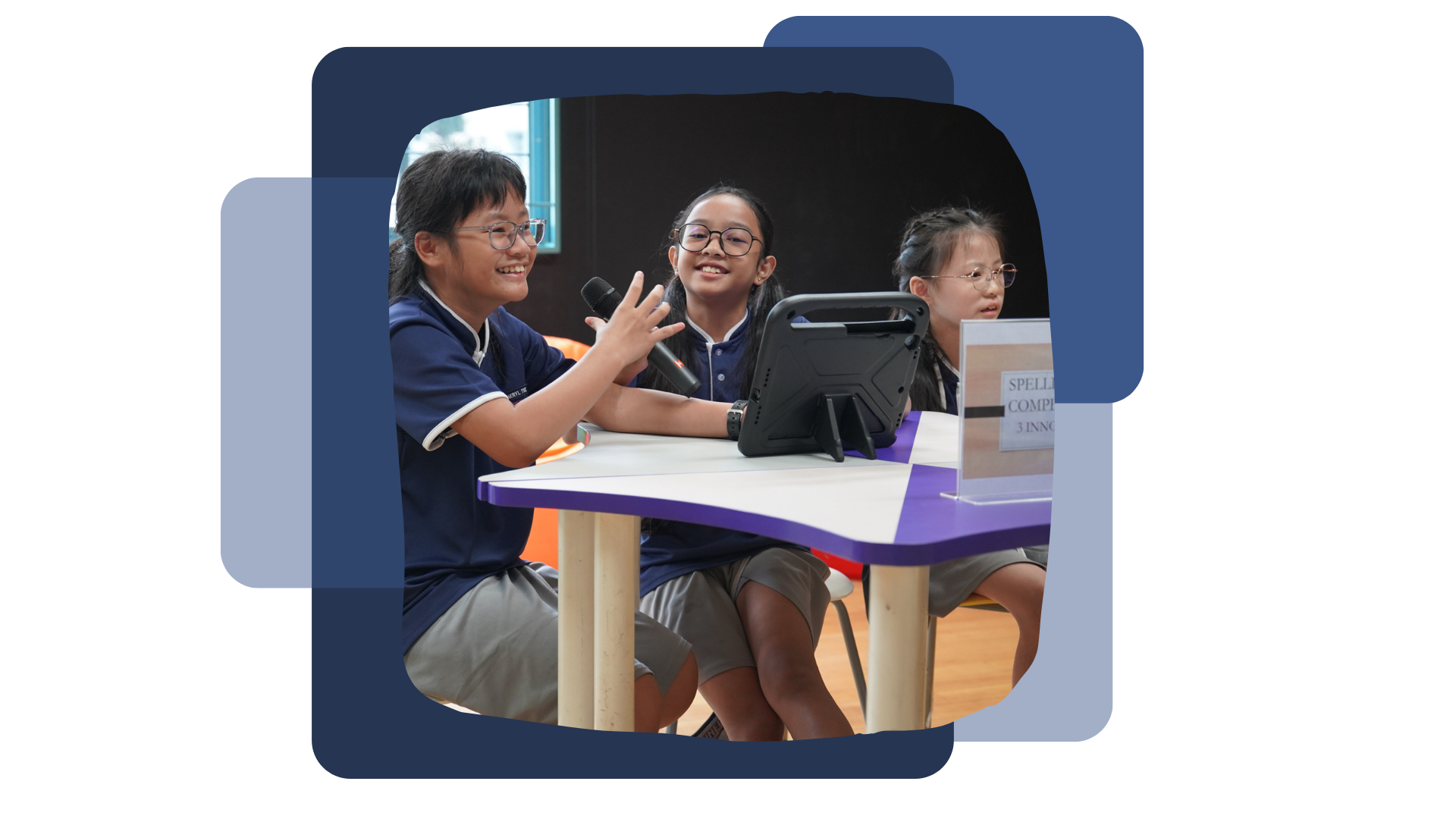 Three smiling school children in uniforms sit at a desk with a tablet and microphone during a spelling competition.
