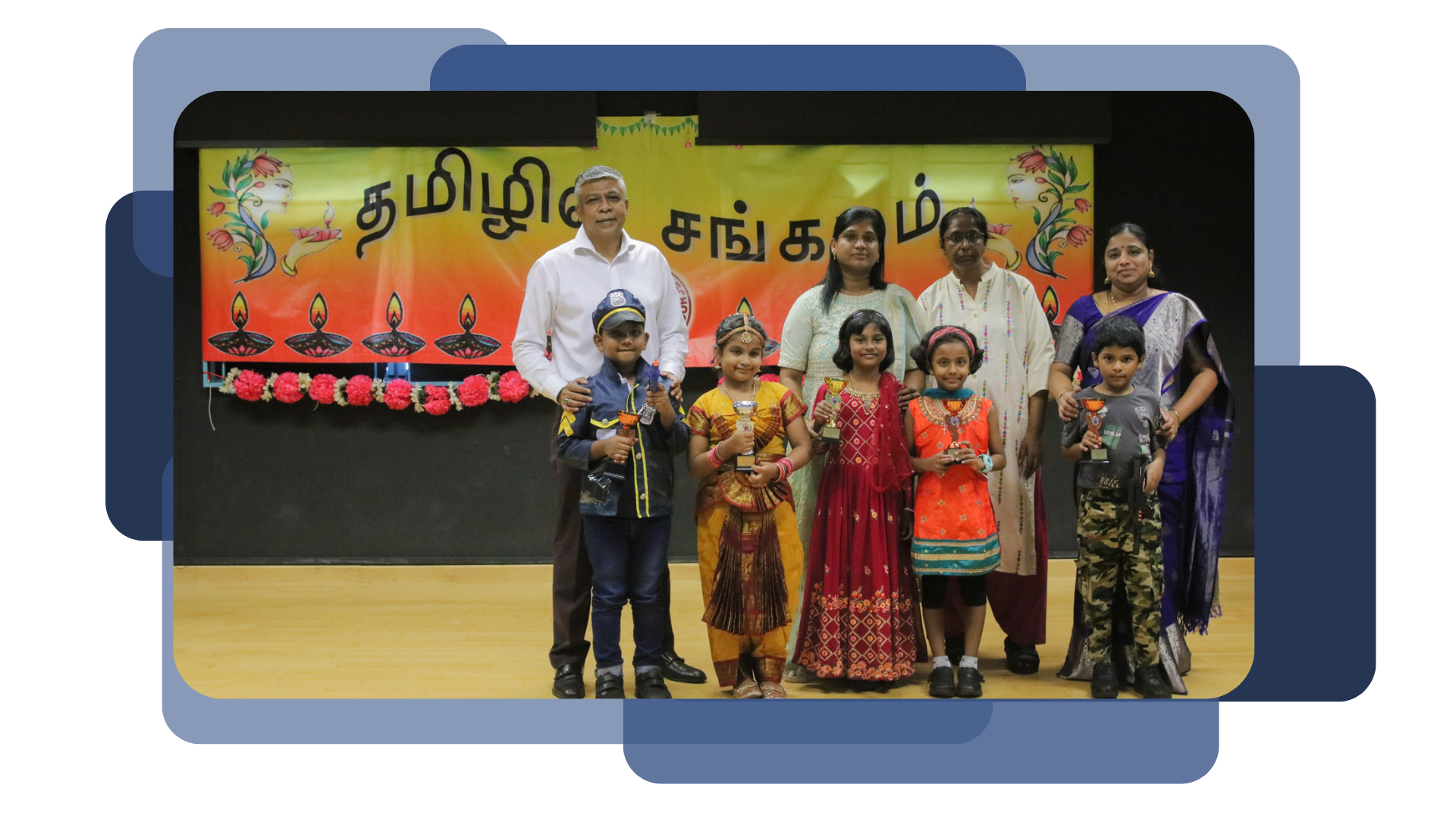 Adults and children in traditional costumes holding trophies on a stage with a Tamil script banner.