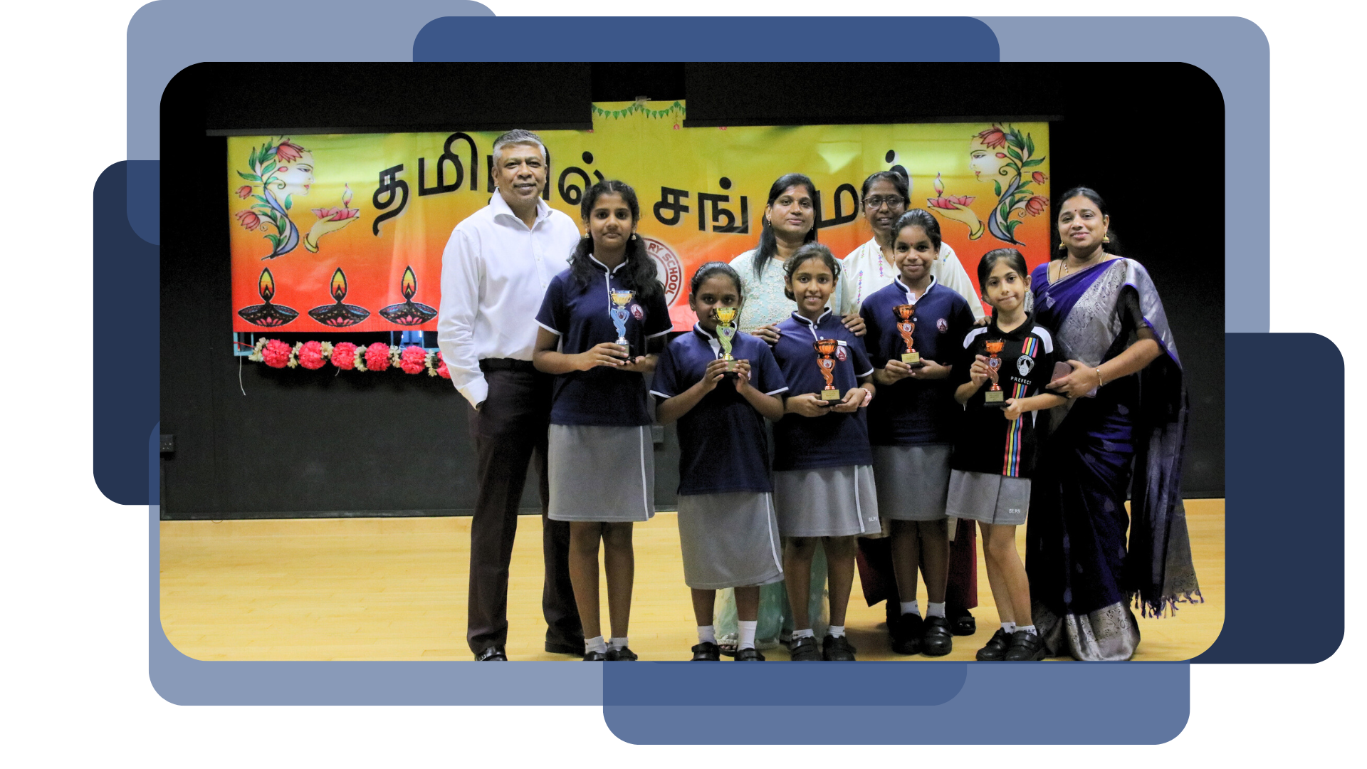 Four girls in school uniforms hold trophies while standing with three adults in front of a Tamil language event banner.