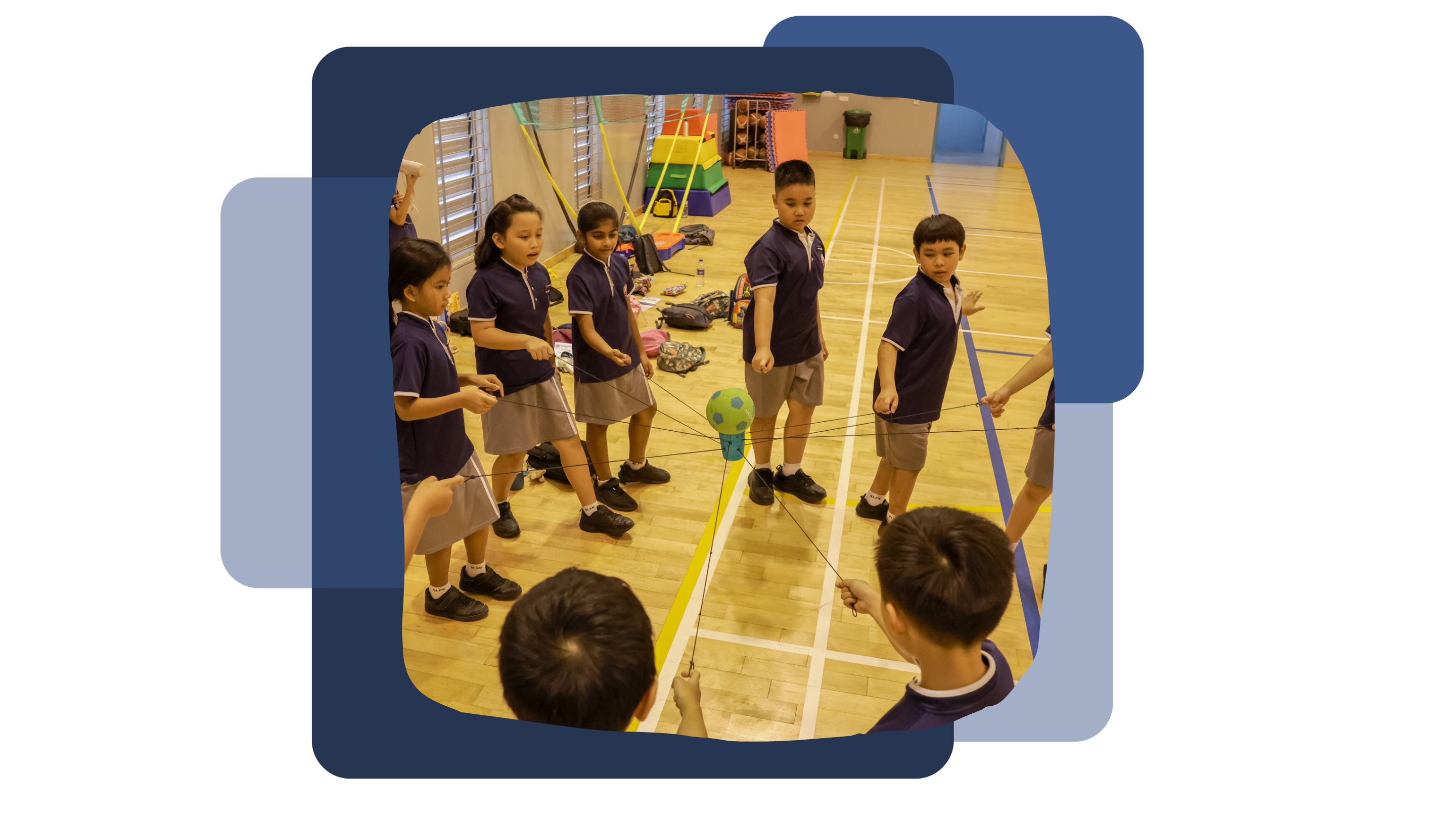 Six children in navy uniforms stand in a circle in a gym, each holding strings attached to a ball.