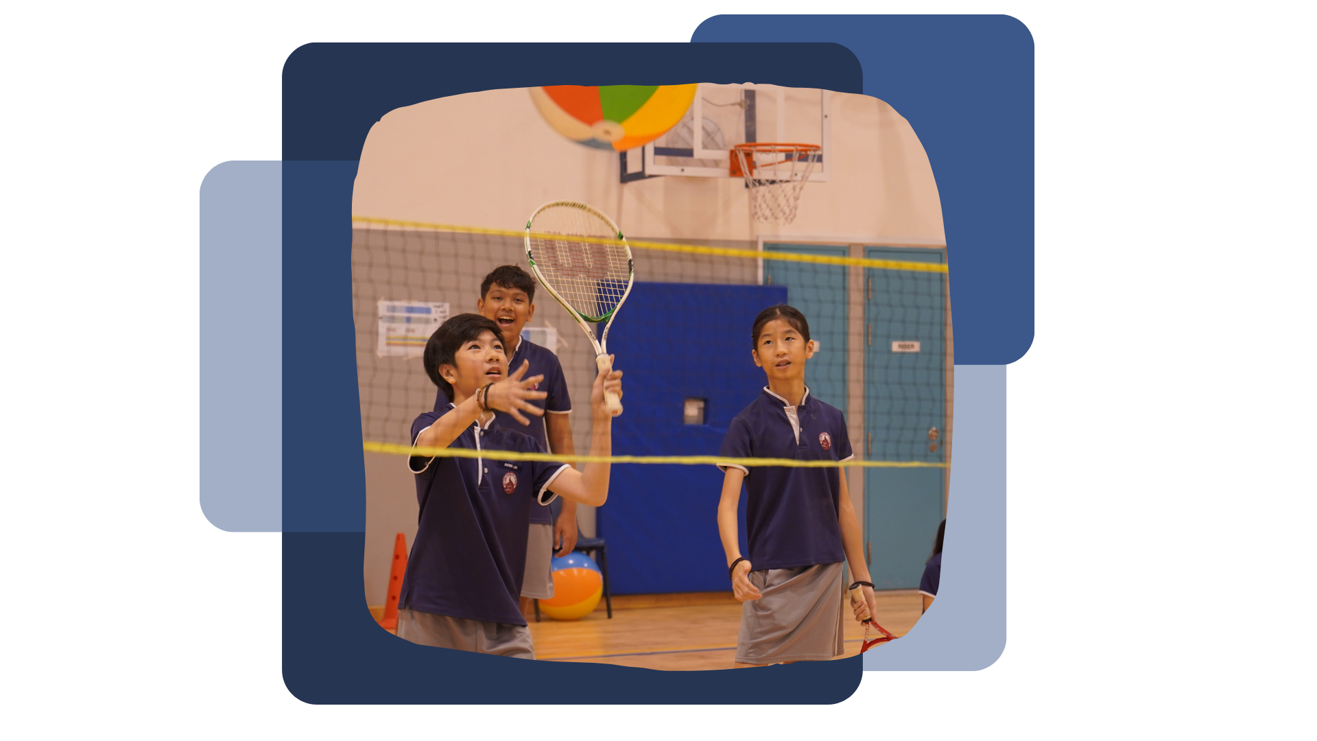Three students in blue uniforms play badminton indoors, one holding a racket up near a net with a basketball hoop behind.