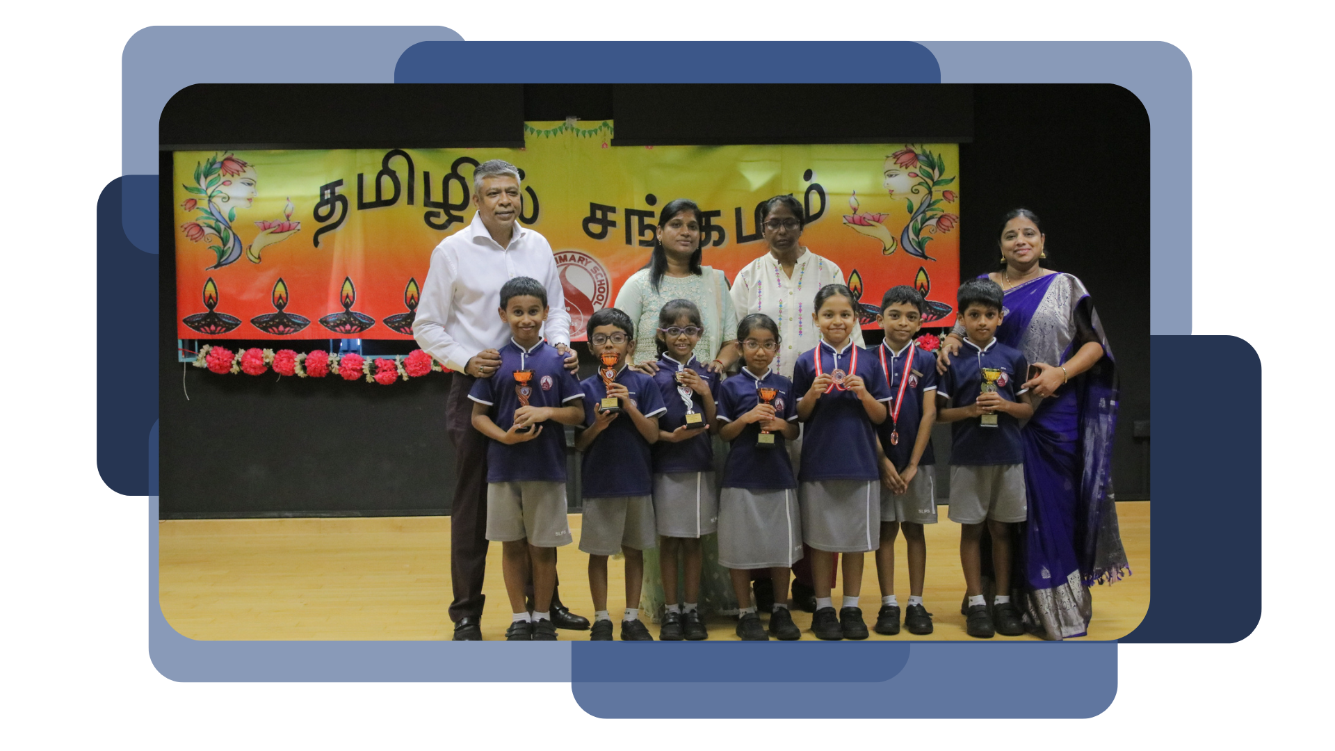 Students in school uniforms hold trophies and medals alongside adults on a stage with a Tamil-script banner.
