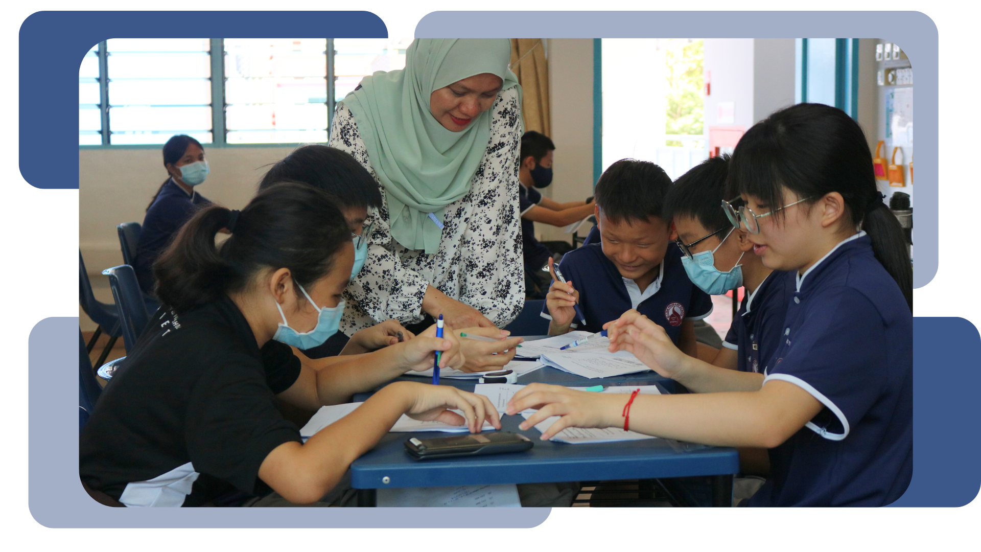A teacher in a green hijab leans over a desk helping masked students with written work in a classroom.