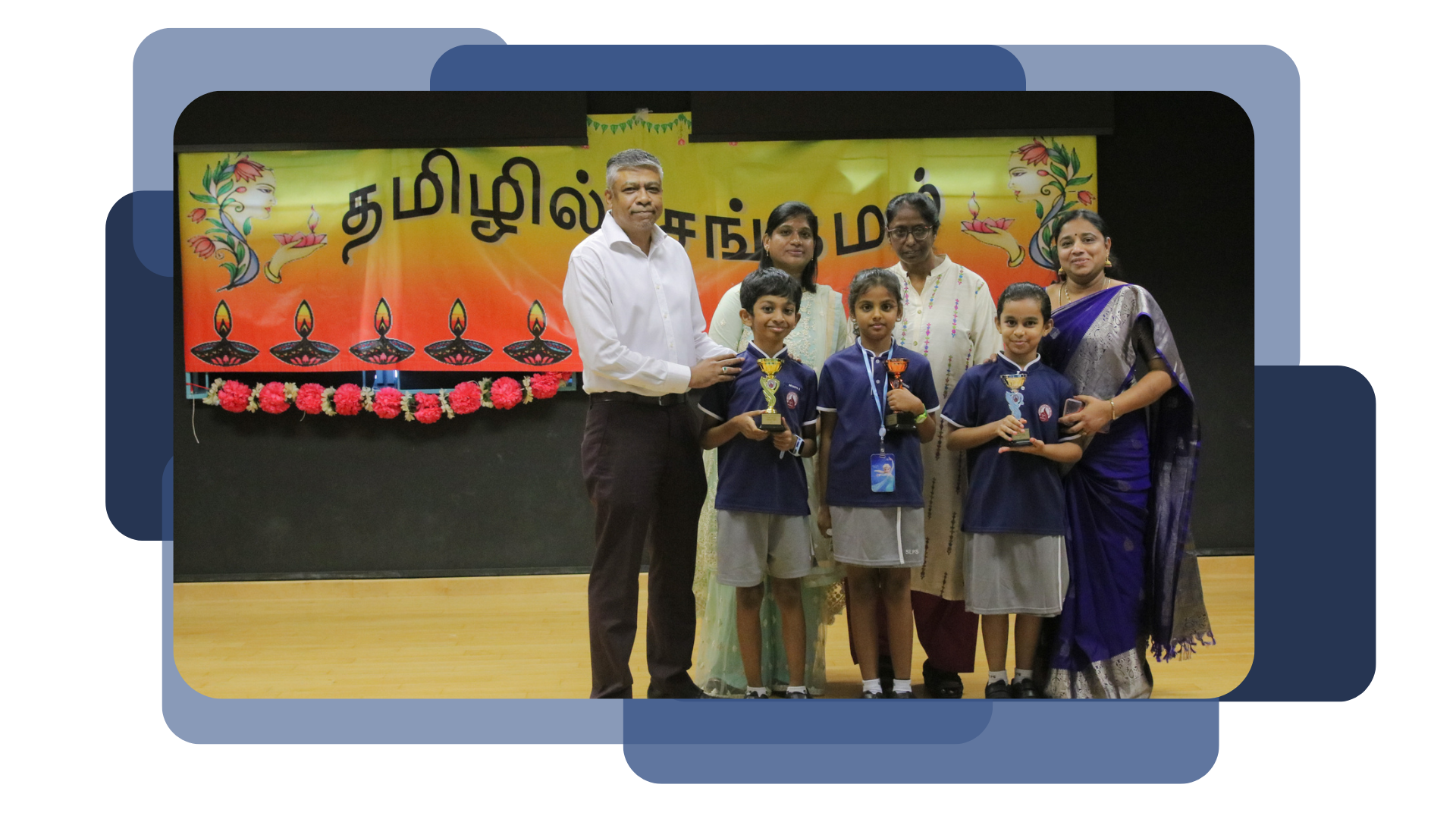 Three children in school uniforms hold trophies on stage with adults before a Tamil-script decorated banner.
