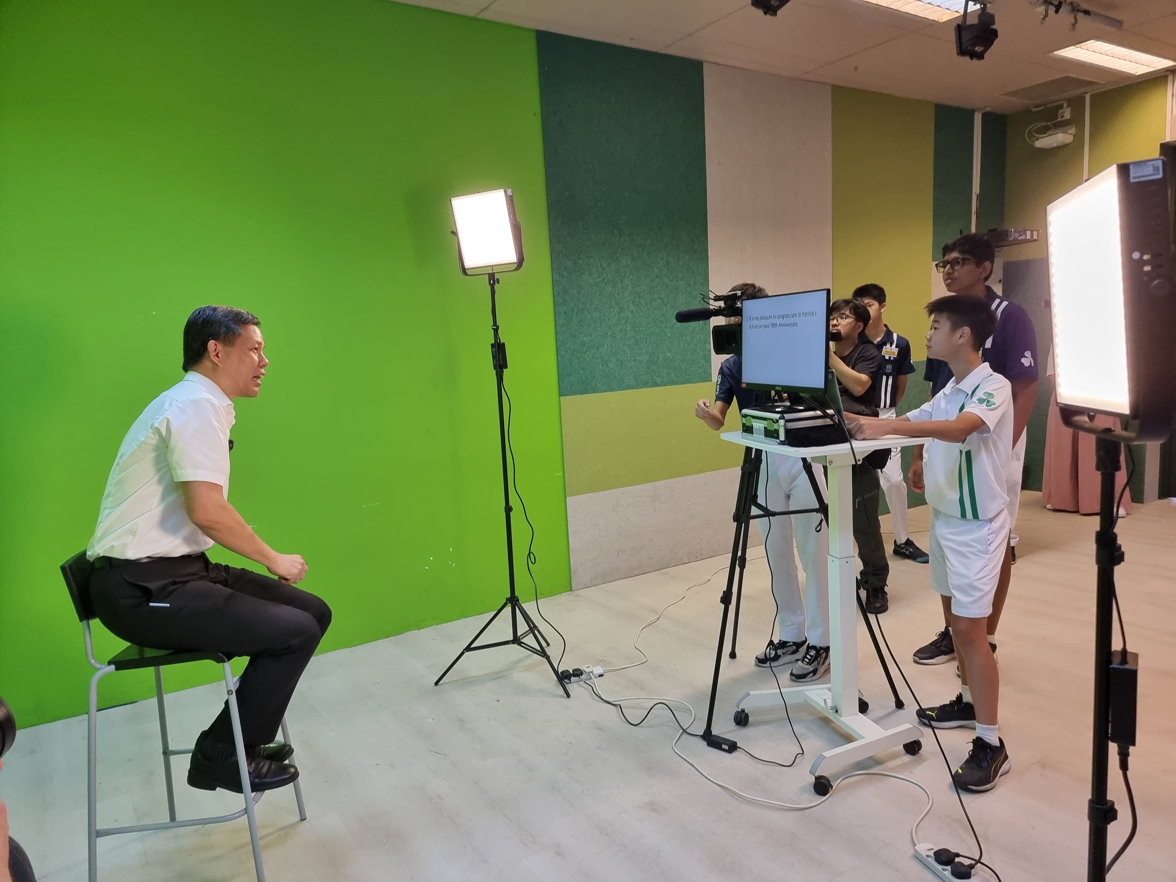 A man sits on a stool before a green screen while three students operate a camera and monitor in a studio.