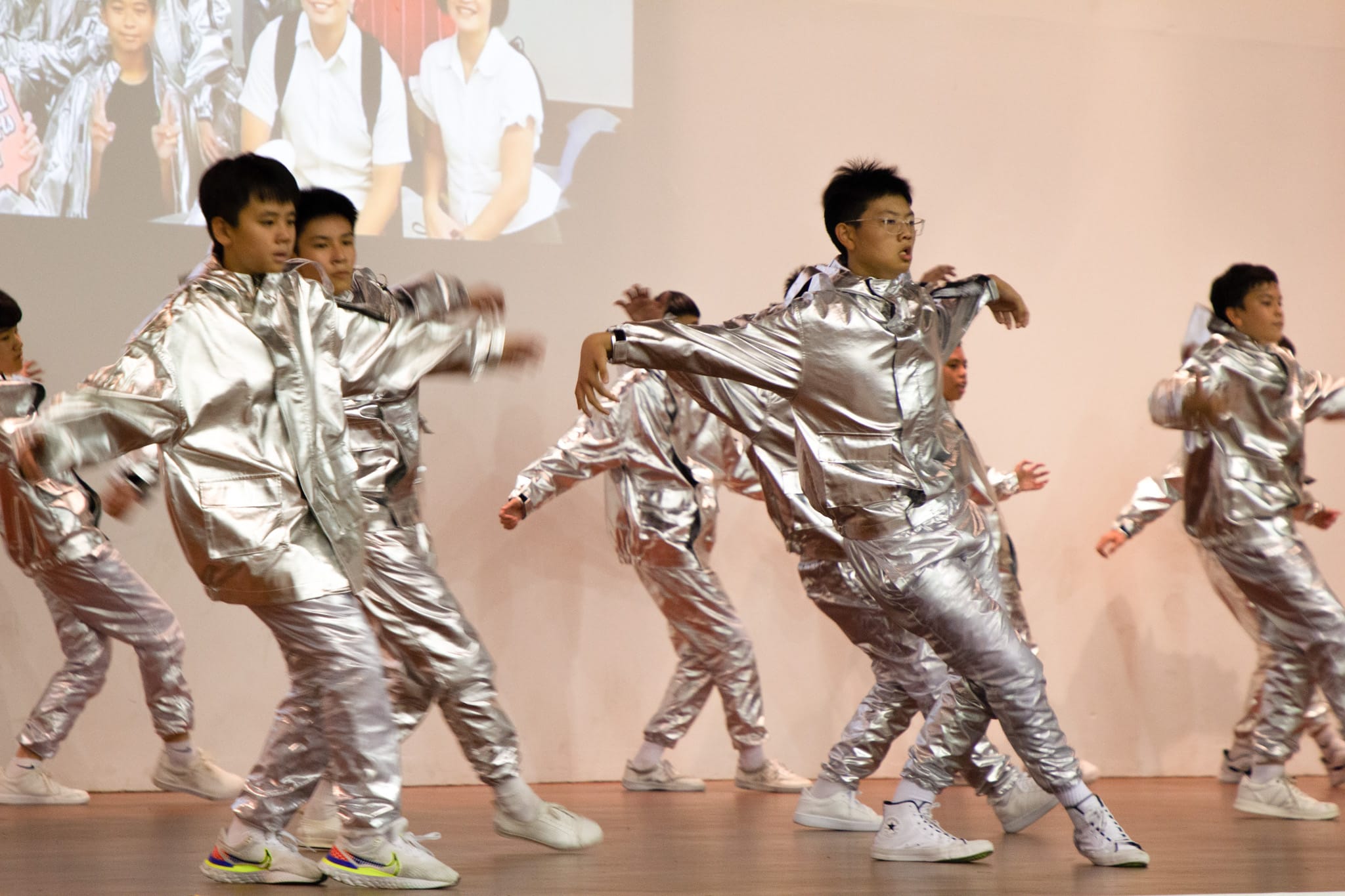 Four young males breakdancing outdoors, one doing a headstand move while others pose around him near a white building.