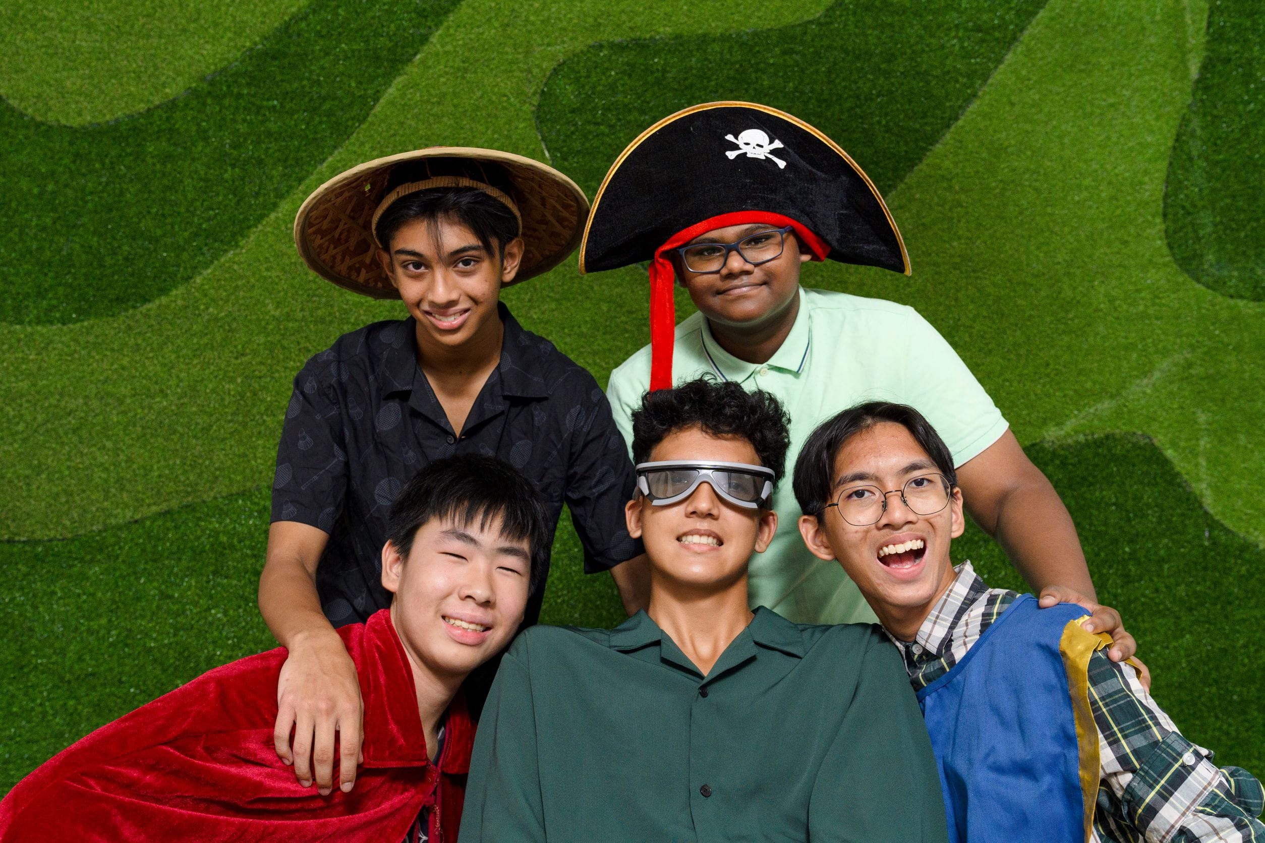 Five teenage boys pose together in costume props including a pirate hat, straw hat, and goggles against a green leafy backdrop.