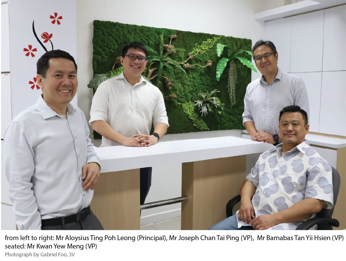 Four men pose in an office with a green moss wall, three standing and one seated, smiling at the camera.