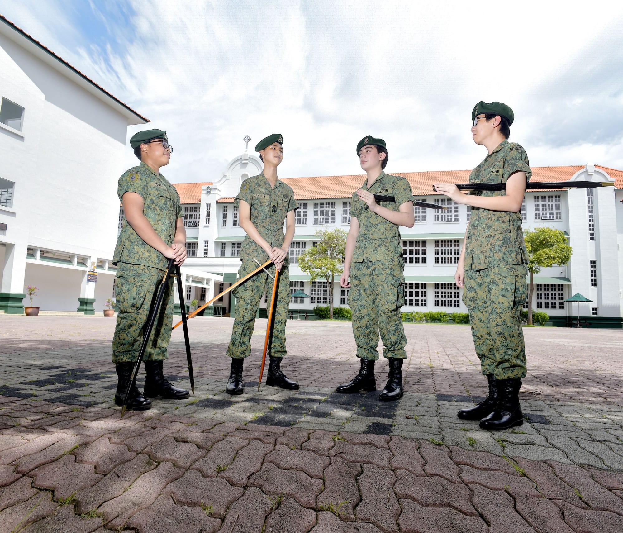 Four soldiers in green camouflage uniforms and berets stand on a paved courtyard, two holding poles, in front of a large building.
