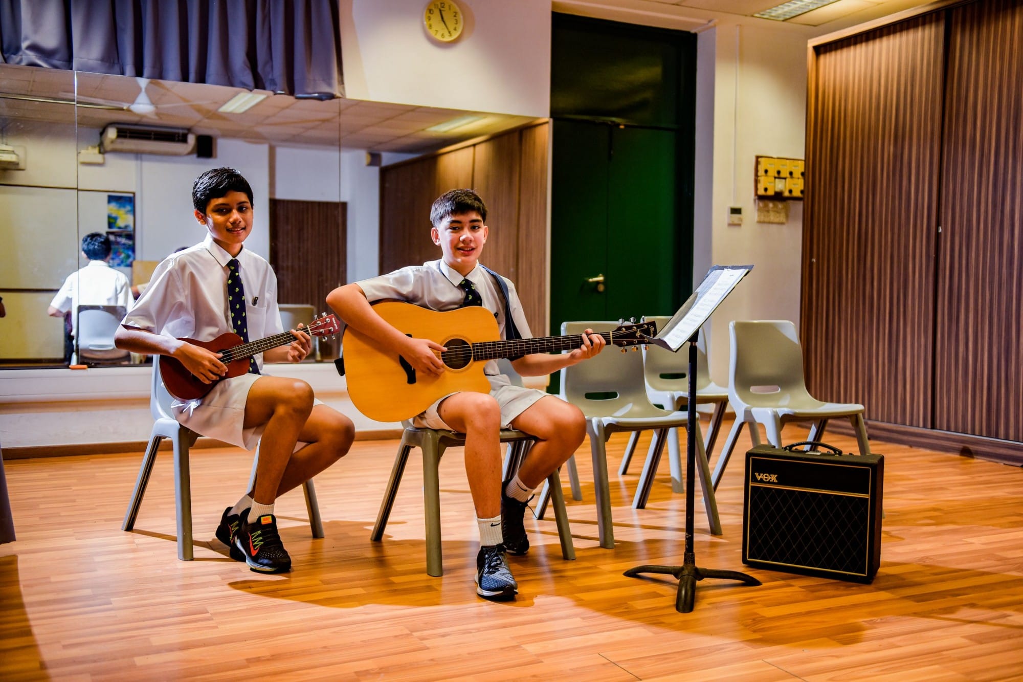 Two school boys in uniforms sit playing a ukulele and acoustic guitar in a music room with a Vox amplifier nearby.