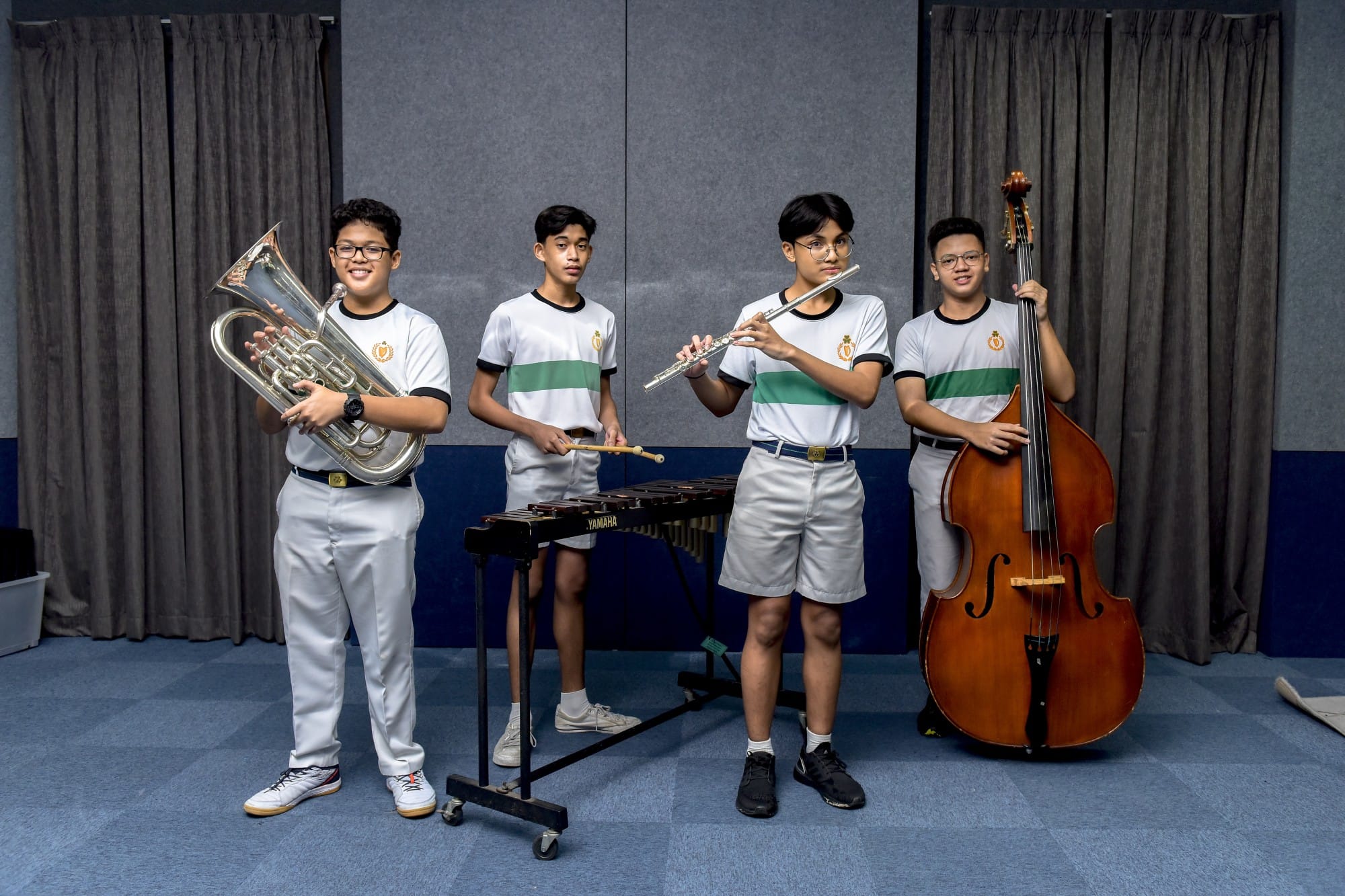 Four students in matching uniforms hold a euphonium, xylophone mallets, flute, and double bass in a music room.