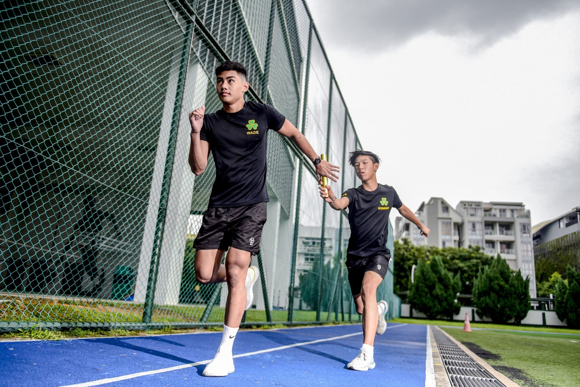 Two young male runners in black uniforms passing a gold baton on a blue athletic track beside a green fence.