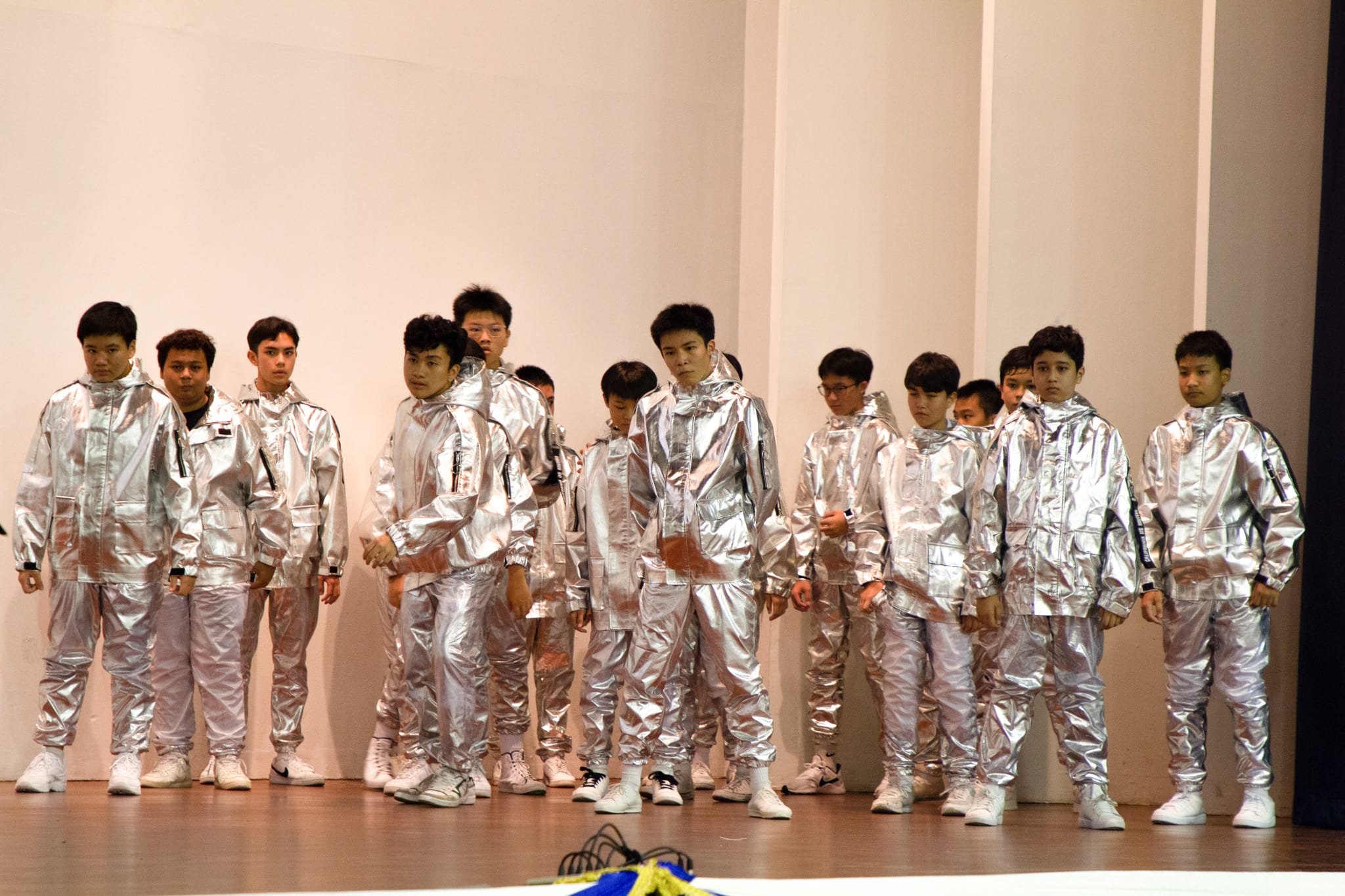 A group of young men standing on a wooden stage, all wearing matching shiny silver jumpsuits and white shoes.