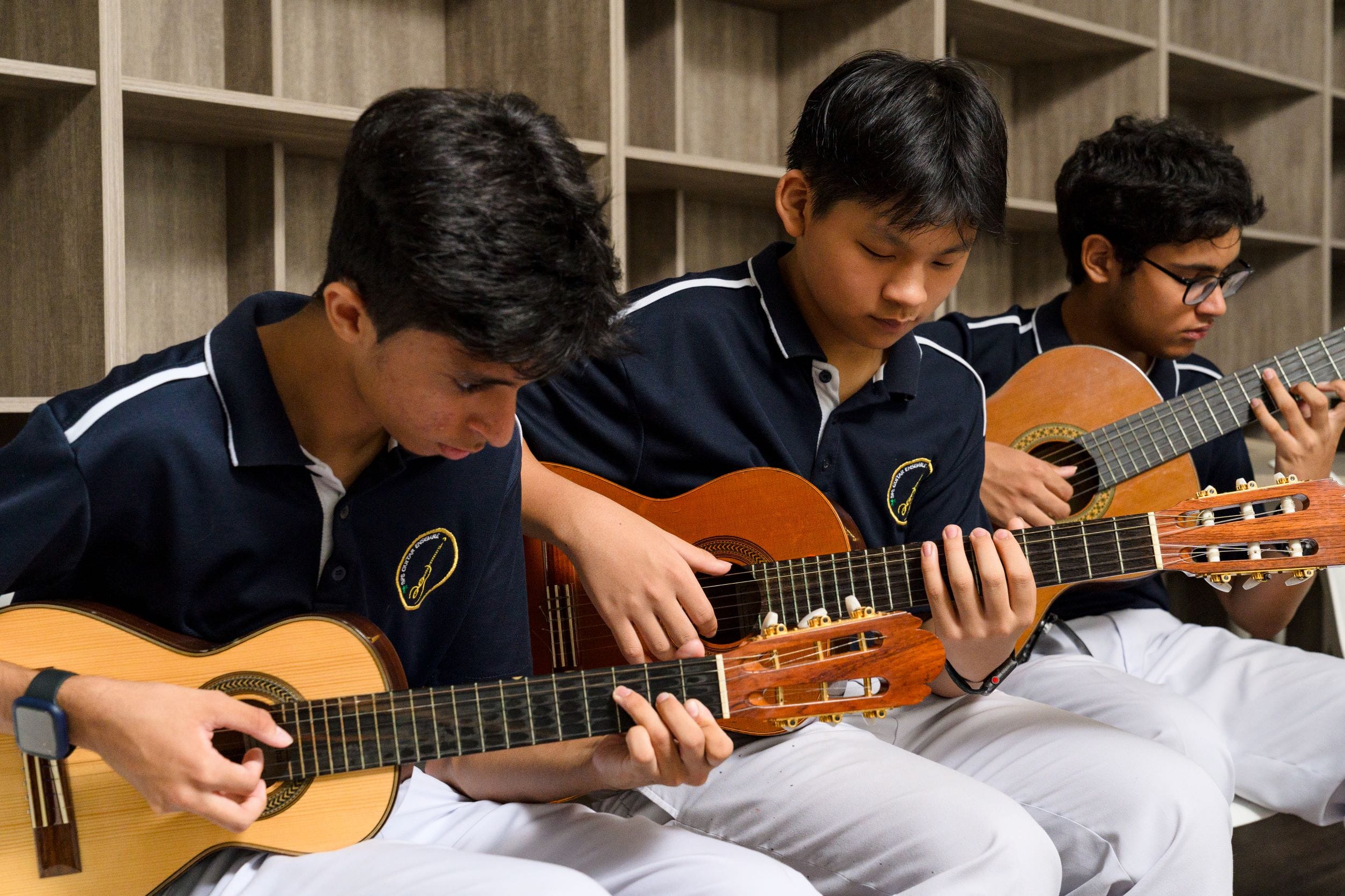 Three teenage boys in navy school uniforms sit together playing acoustic guitars indoors.