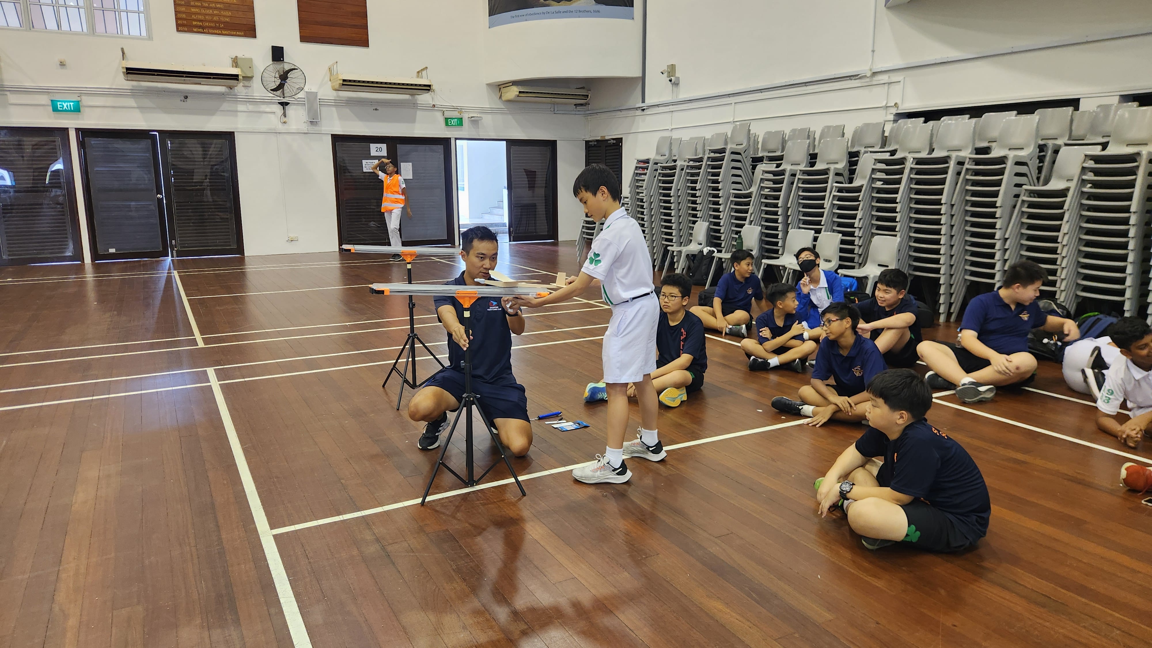 A student hands material to an adult kneeling by a launcher on a wooden indoor court, with other students seated watching.