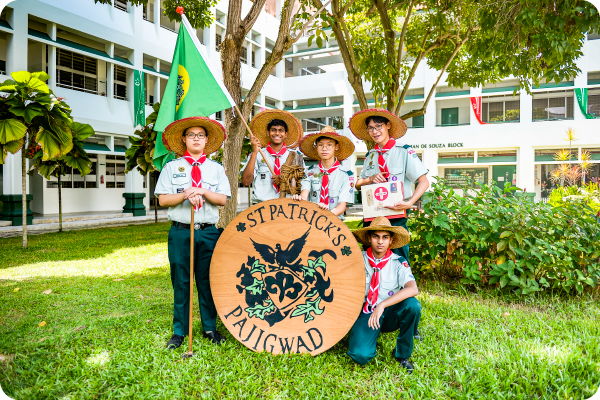 Five scouts in straw hats pose with a St. Patrick's Patrol sign and green flag outside a school building.