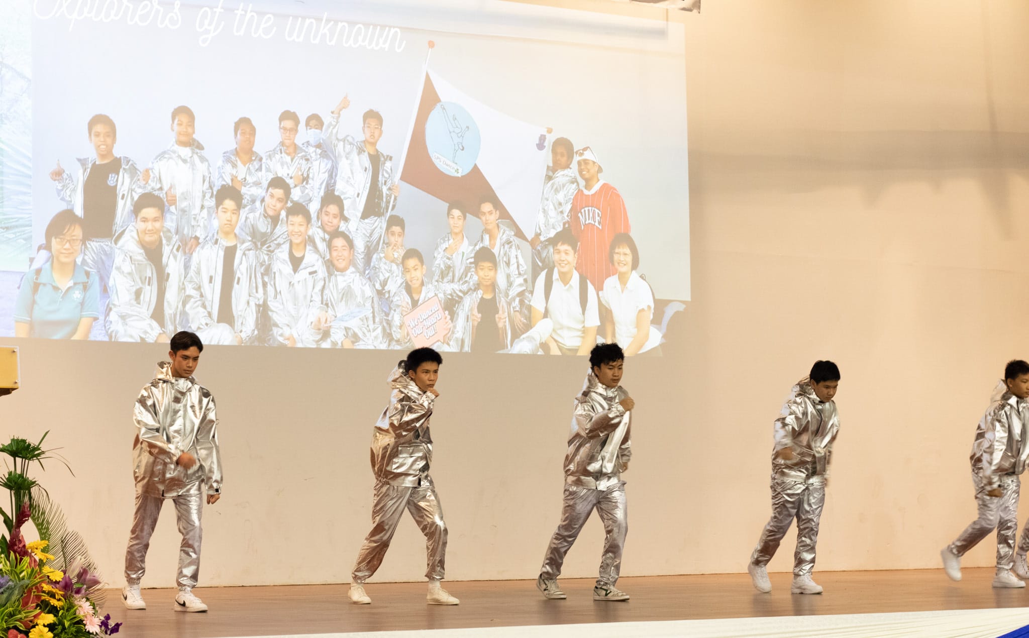 Five young male performers in silver metallic suits dance on stage with a projected group photo behind them.