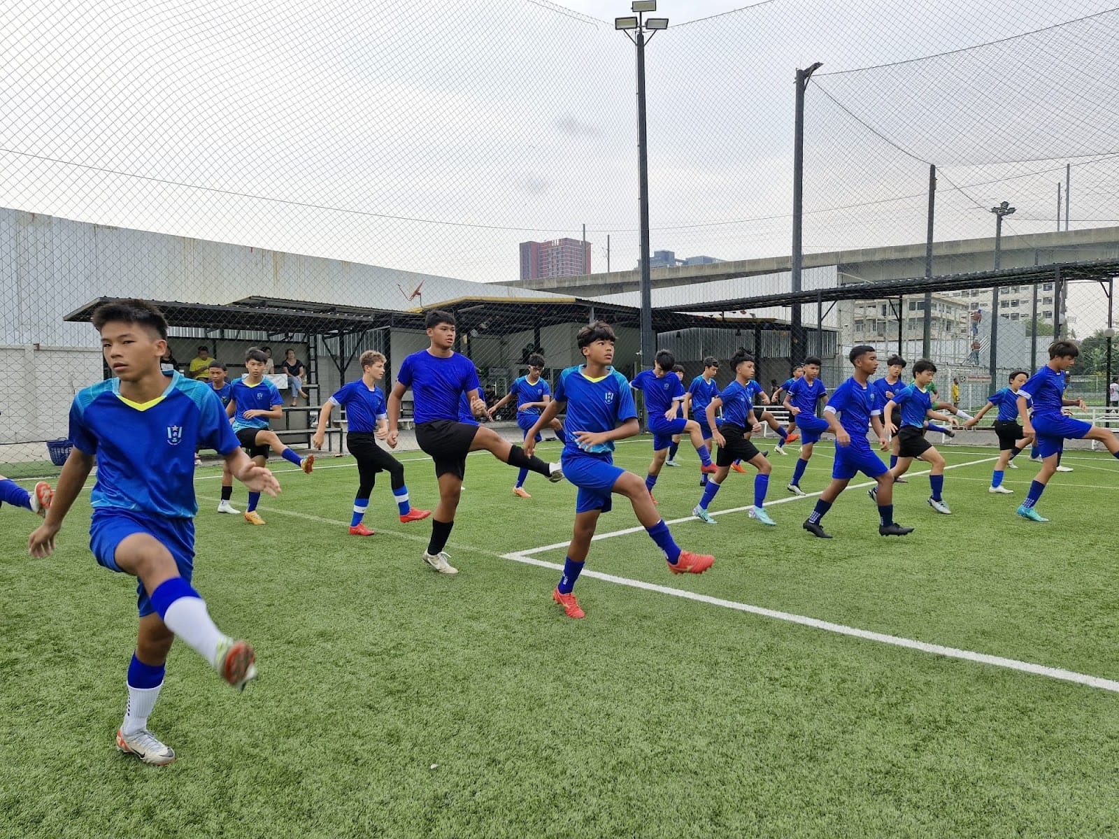A large group of boys in blue soccer uniforms doing warm-up drills on a green turf field.