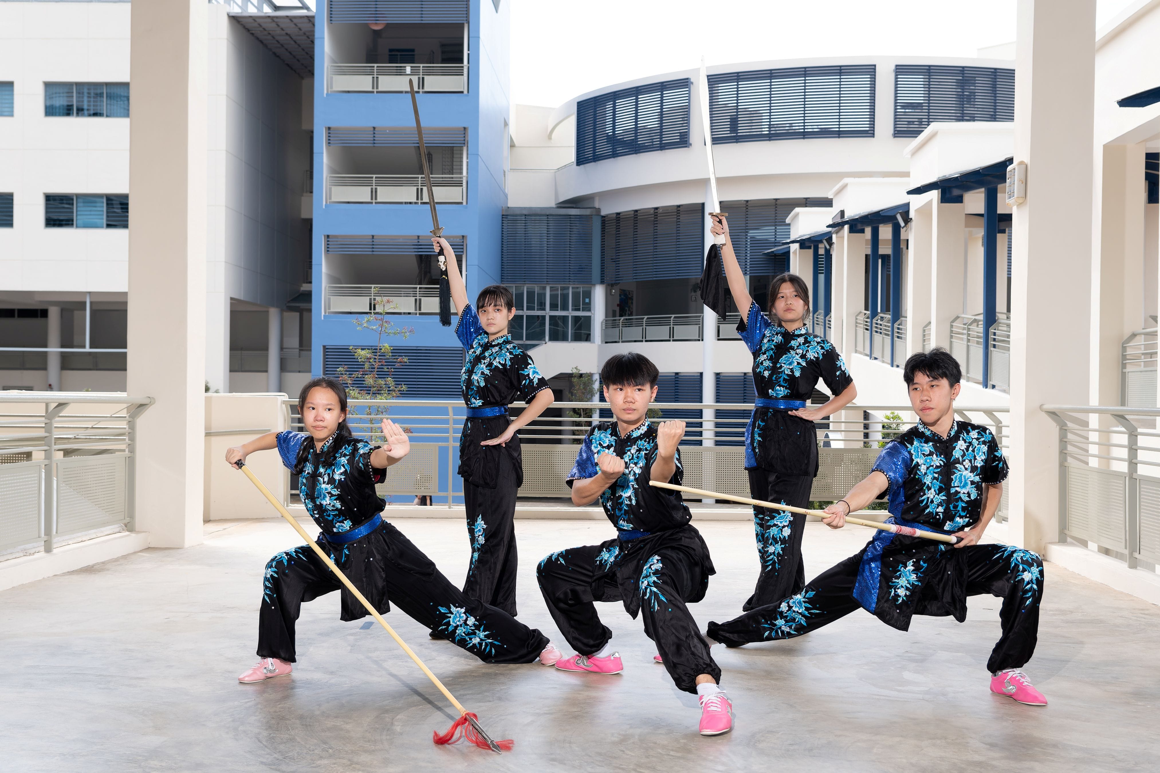 Five TMJC wushu students in matching black and blue floral uniforms strike dynamic martial arts poses outdoors, holding weapons including staffs and swords.