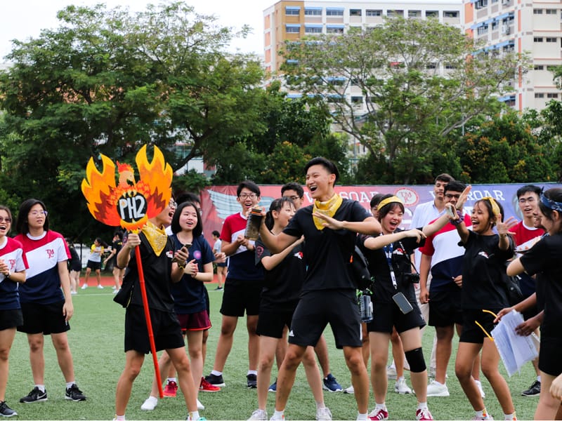 Students celebrate on a green field, one holding a flame-shaped "PH2" sign, with buildings and trees behind them.