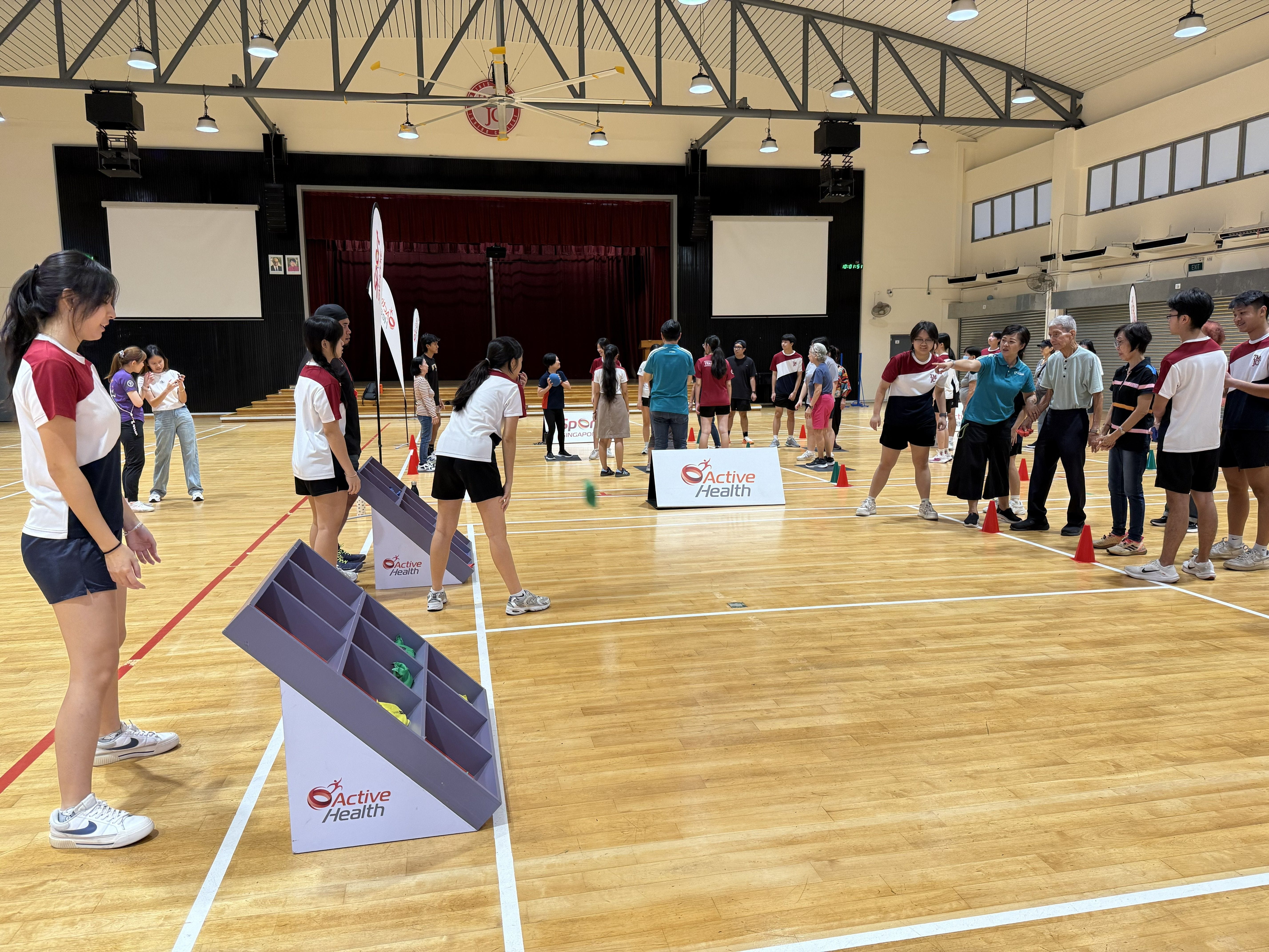 Students and elderly participants engage in an Active Health sports activity inside a school hall. They are playing a target-tossing game using an angled rack, with Active Health and Sport Singapore branding visible throughout.