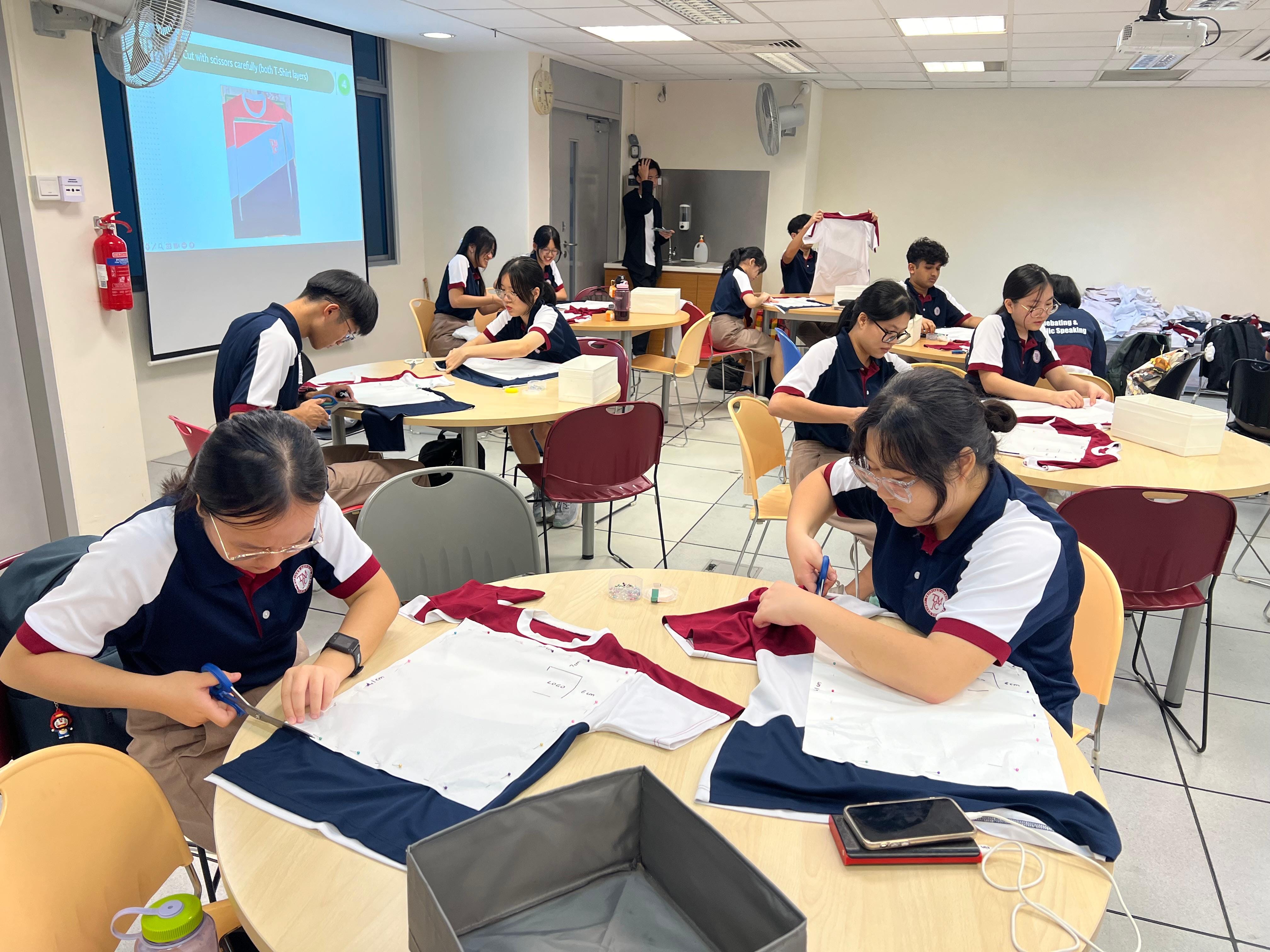 Students in school uniforms sit at round tables in a classroom, cutting and marking fabric pieces. A projected screen at the front displays instructions for cutting a T-shirt.