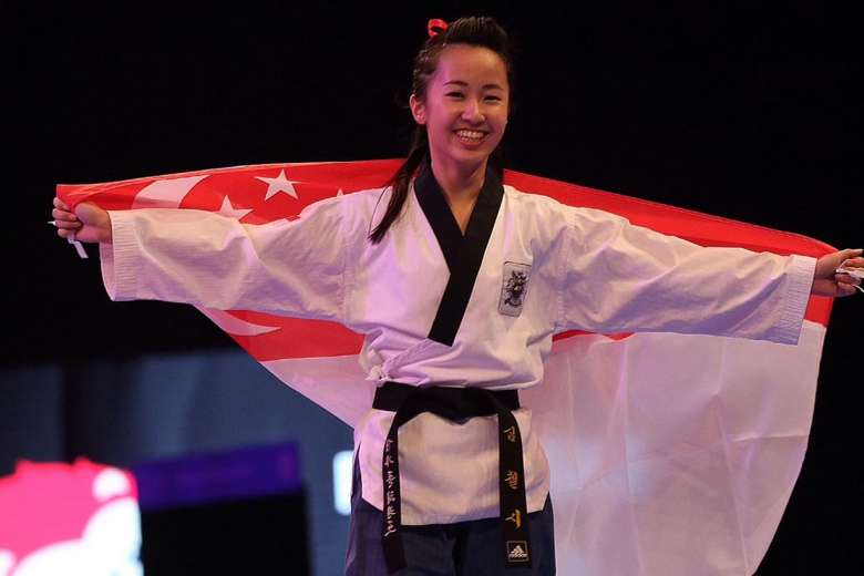 Smiling female taekwondo athlete in a white uniform with black belt holds the Singapore flag wide open.
